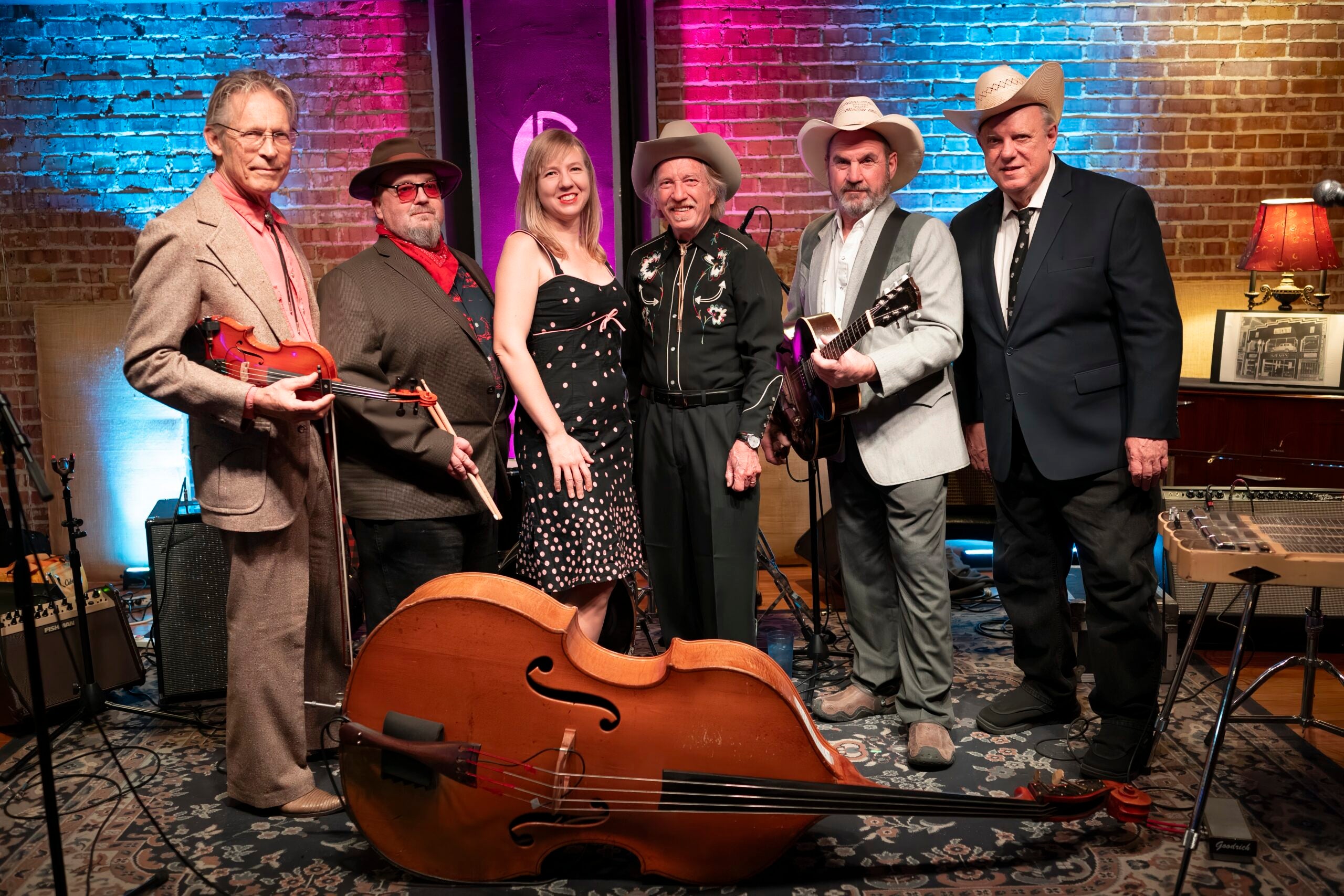 Six musicians, four men and one woman, stand together with instruments including a violin, guitar, and upright bass, in front of a brick wall with stage lighting.