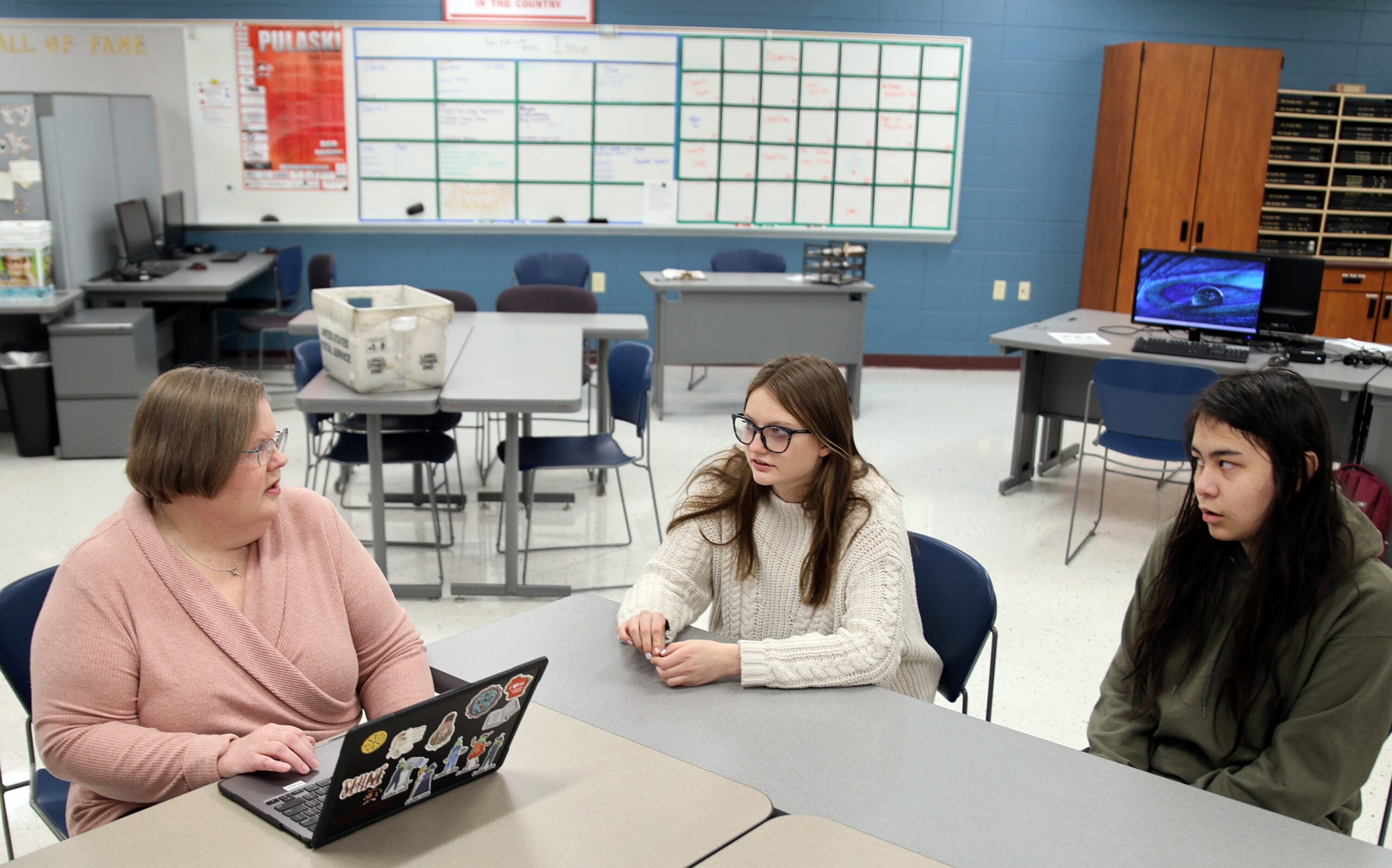 Three women sit around a table in a classroom with computers, desks, and a whiteboard in the background, engaged in conversation.