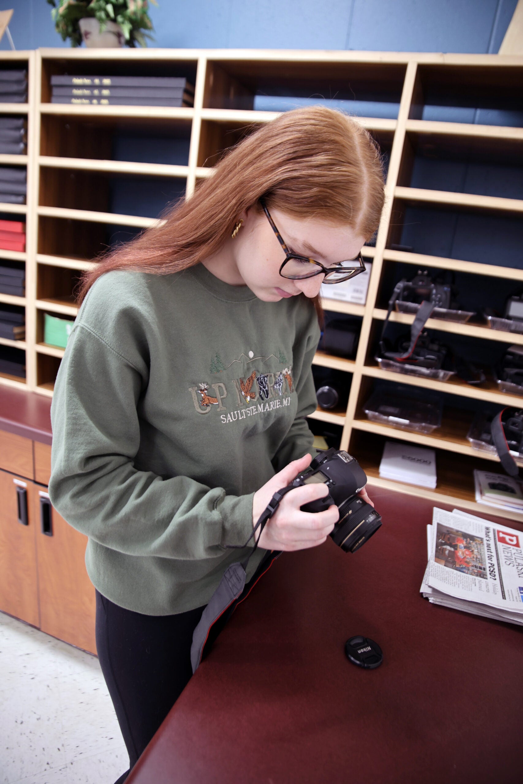 A person with long red hair and glasses examines a camera in front of wood cubby shelves and a red counter with a newspaper and lens cap.