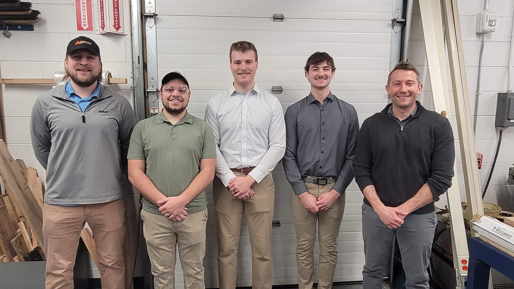 Five men standing indoors in front of a closed white garage door, all facing the camera and smiling.