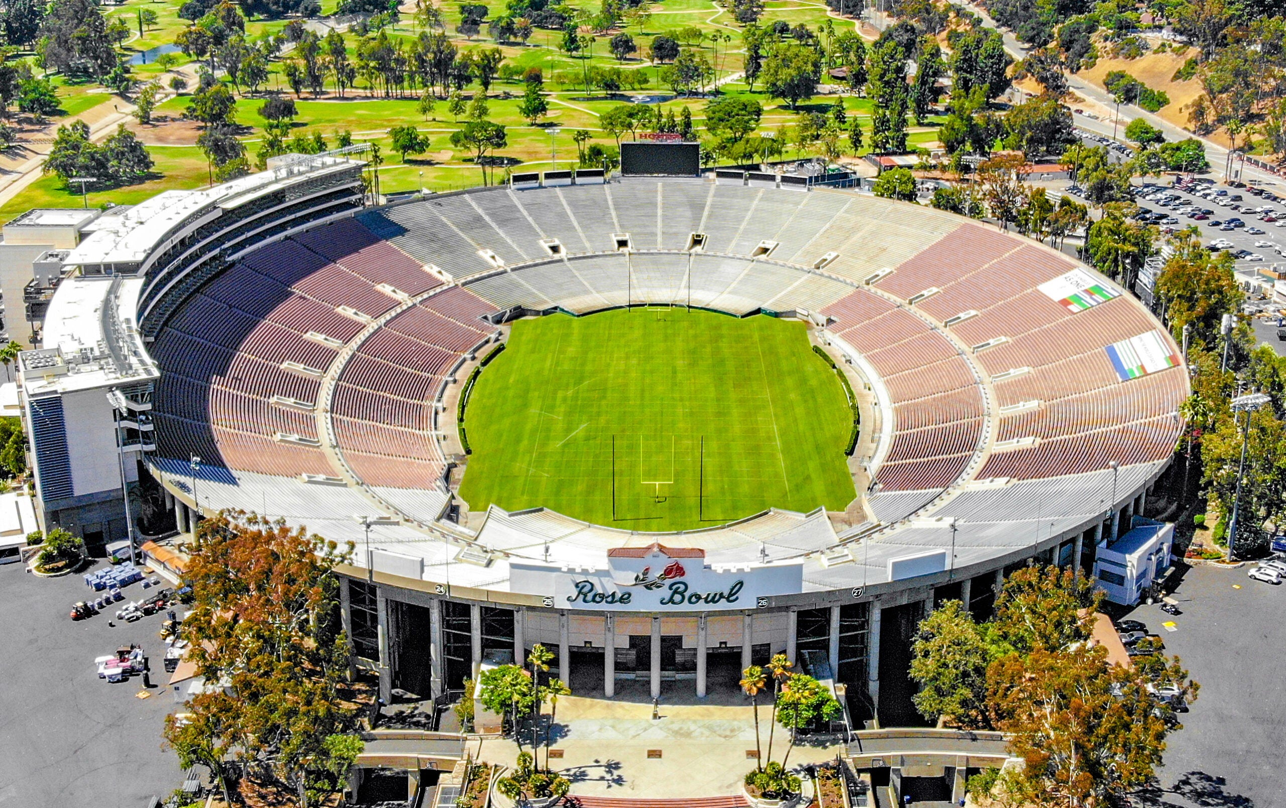 Aerial view of the Rose Bowl stadium showing the empty seating, green field, and surrounding trees and parking areas on a sunny day.