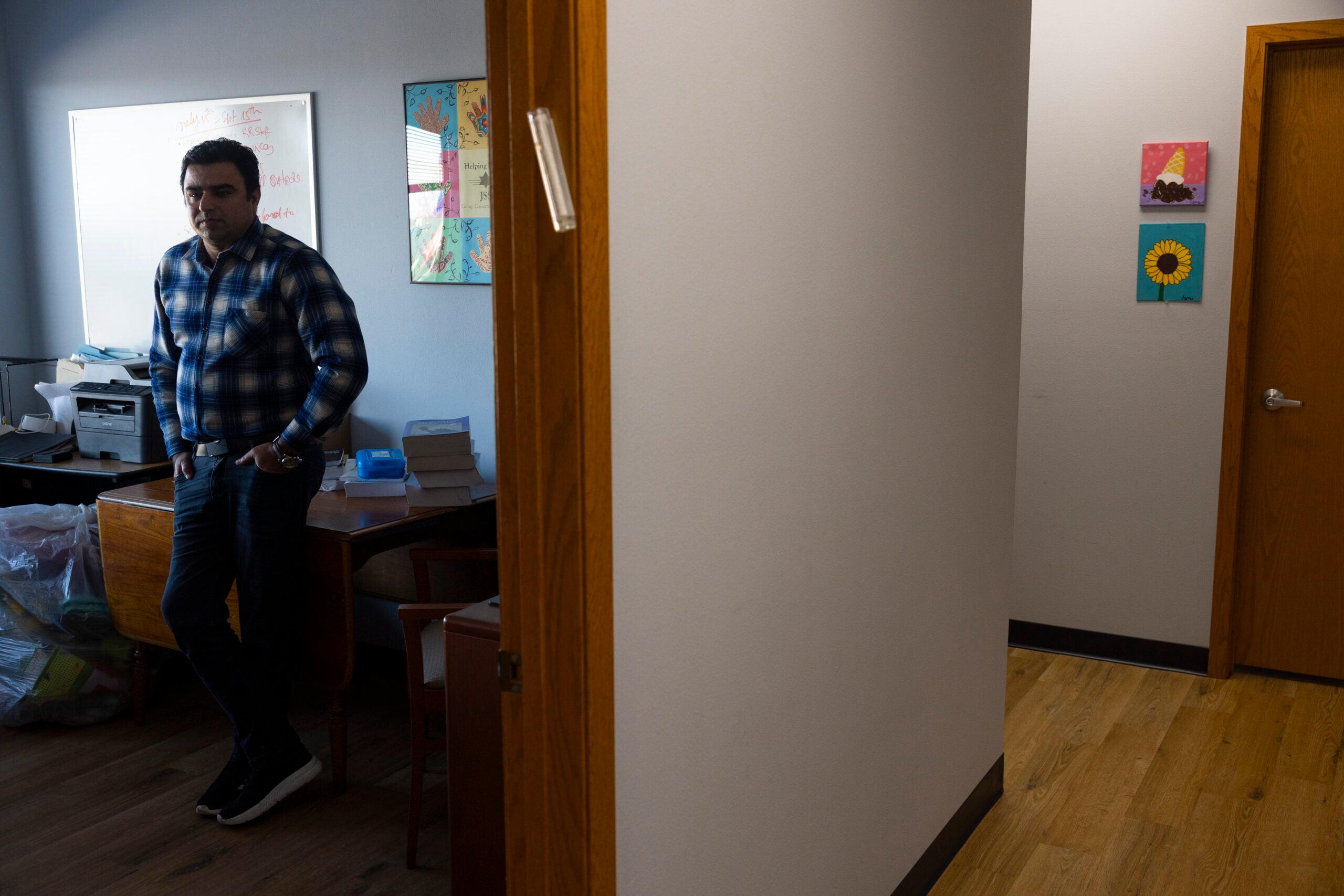 A man in a plaid shirt stands in an office near a desk with papers and bags; a hallway with art on the walls is visible to the right.