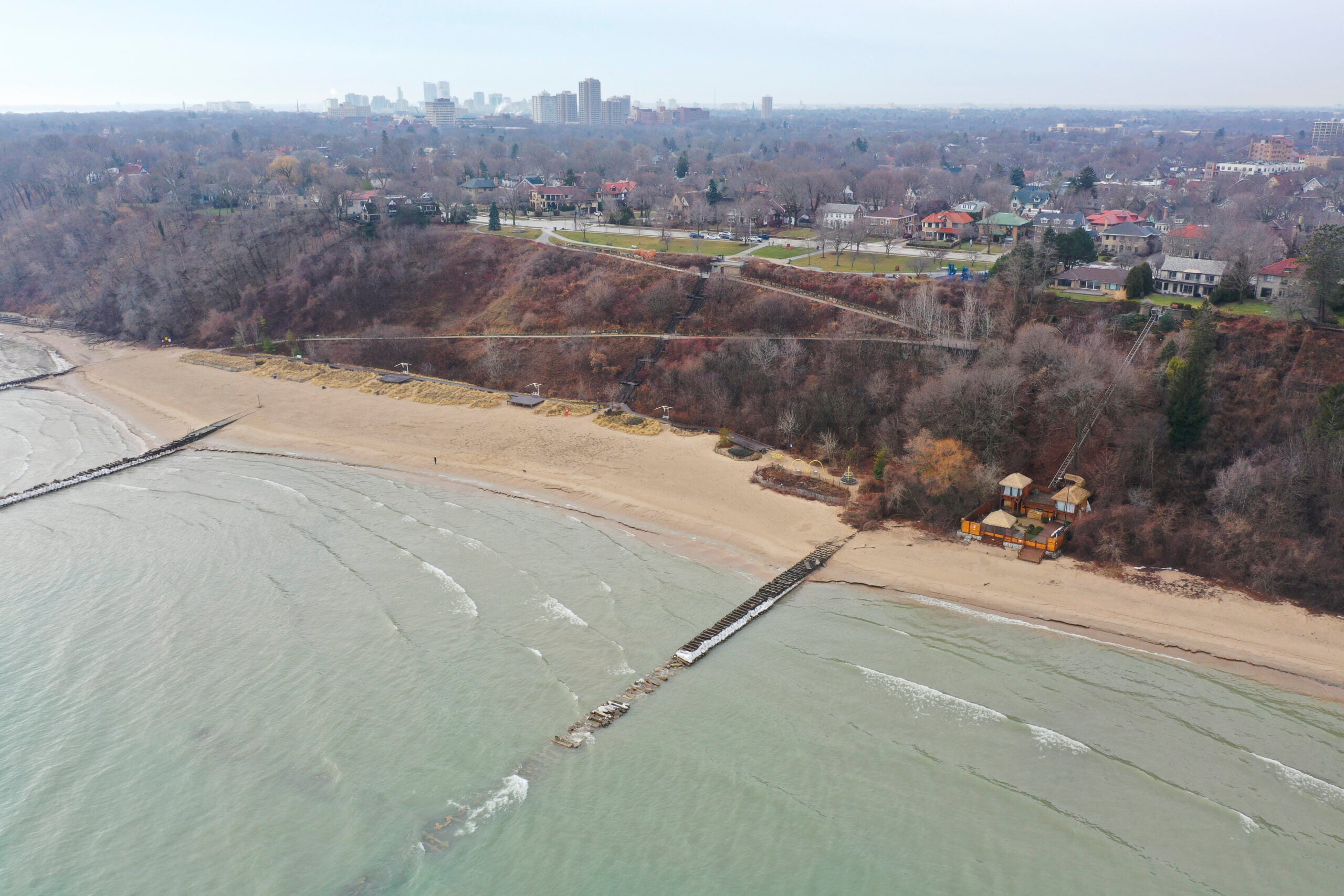 Aerial view of a sandy beach with wave breakers, adjacent to a tree-covered bluff and residential area, with a city skyline in the distance.