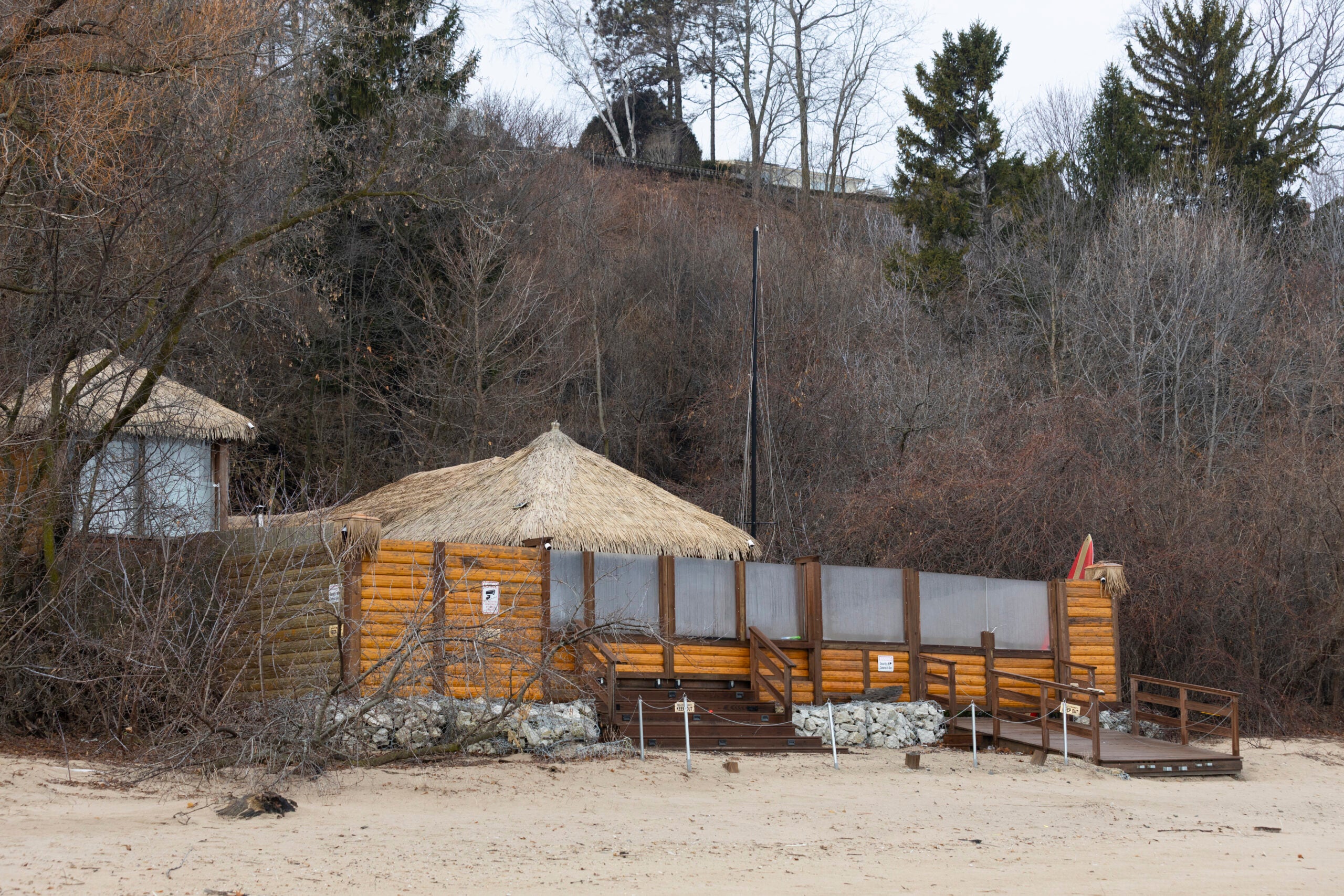 A closed wooden beach hut with a thatched roof sits on sandy ground, surrounded by leafless trees and brush on a cloudy day.