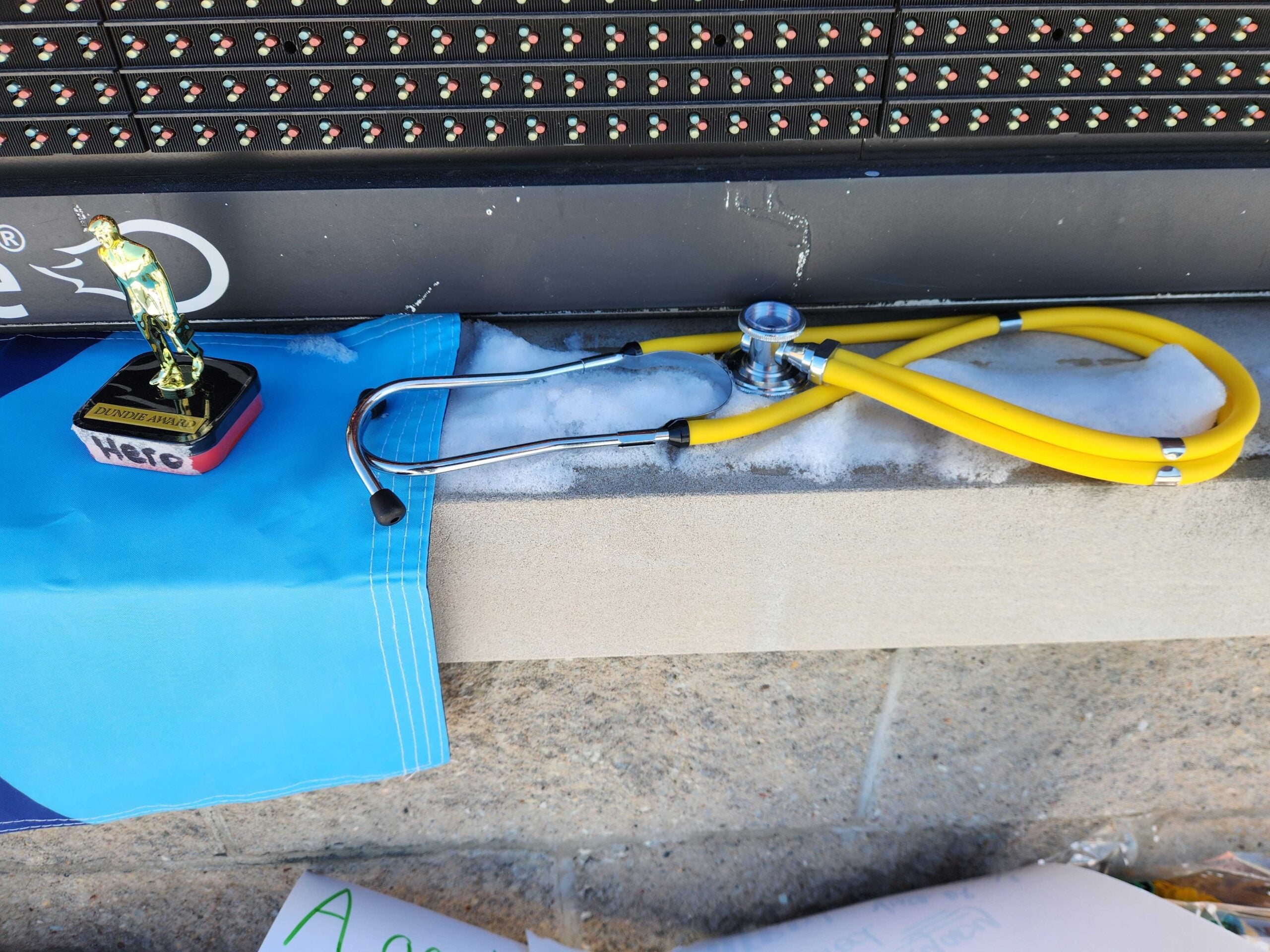 A yellow stethoscope, a small Hero trophy, and a blue cloth rest on a snowy concrete ledge in front of an electronic sign.