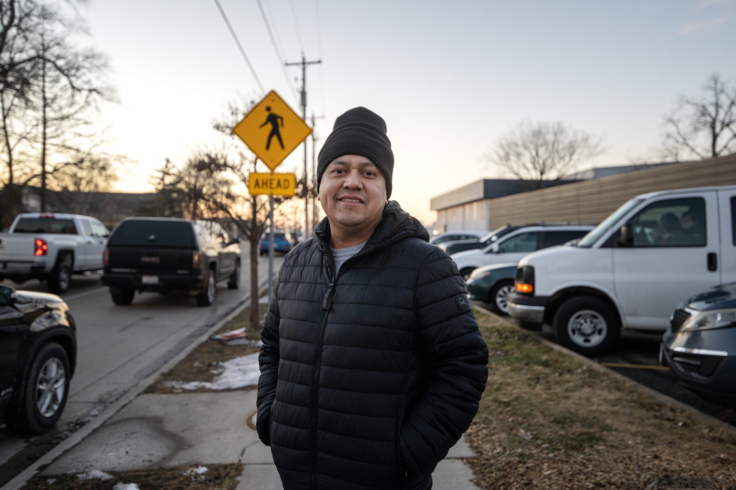 A man wearing a black beanie and puffer jacket stands on a sidewalk near a busy street with cars and a pedestrian crossing sign in the background.