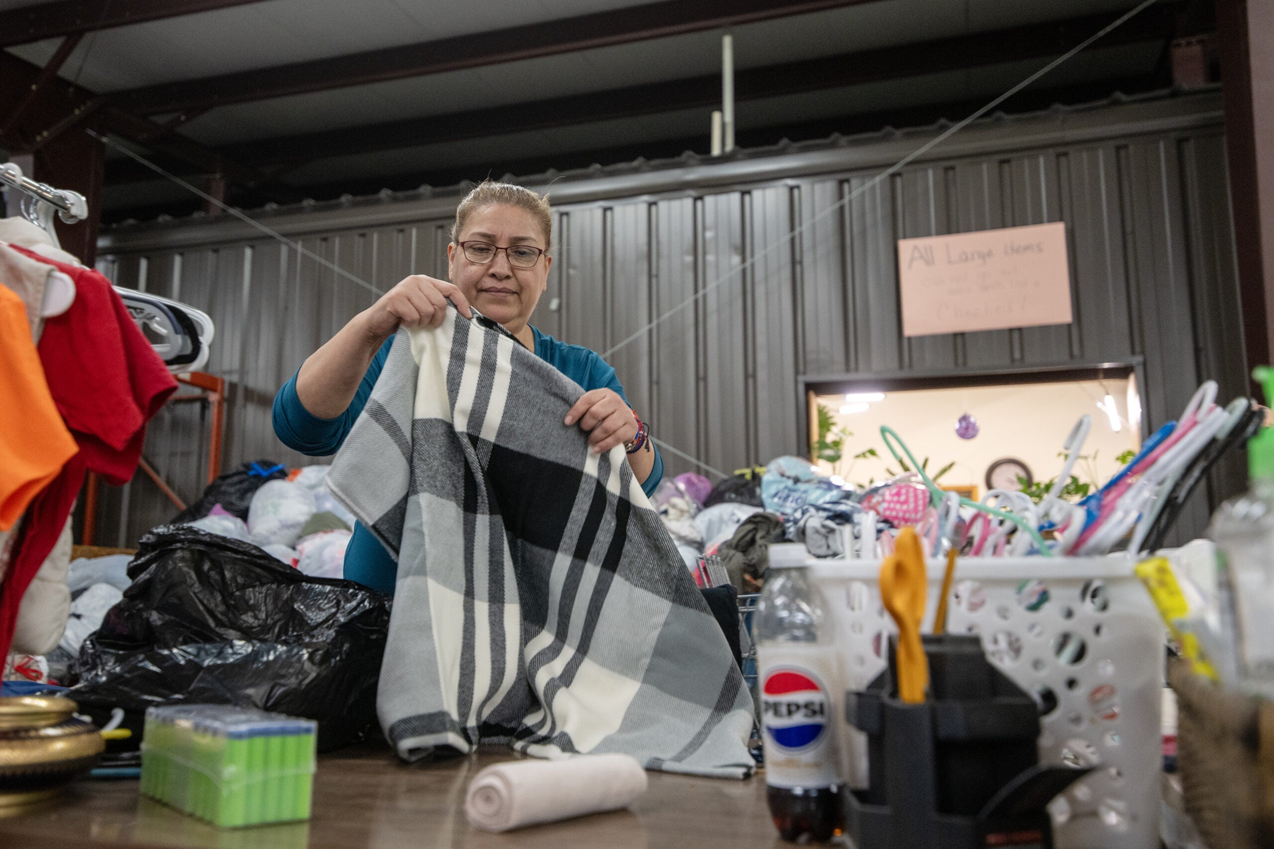 A woman folds a plaid blanket at a table filled with donated items in what appears to be a donation center.