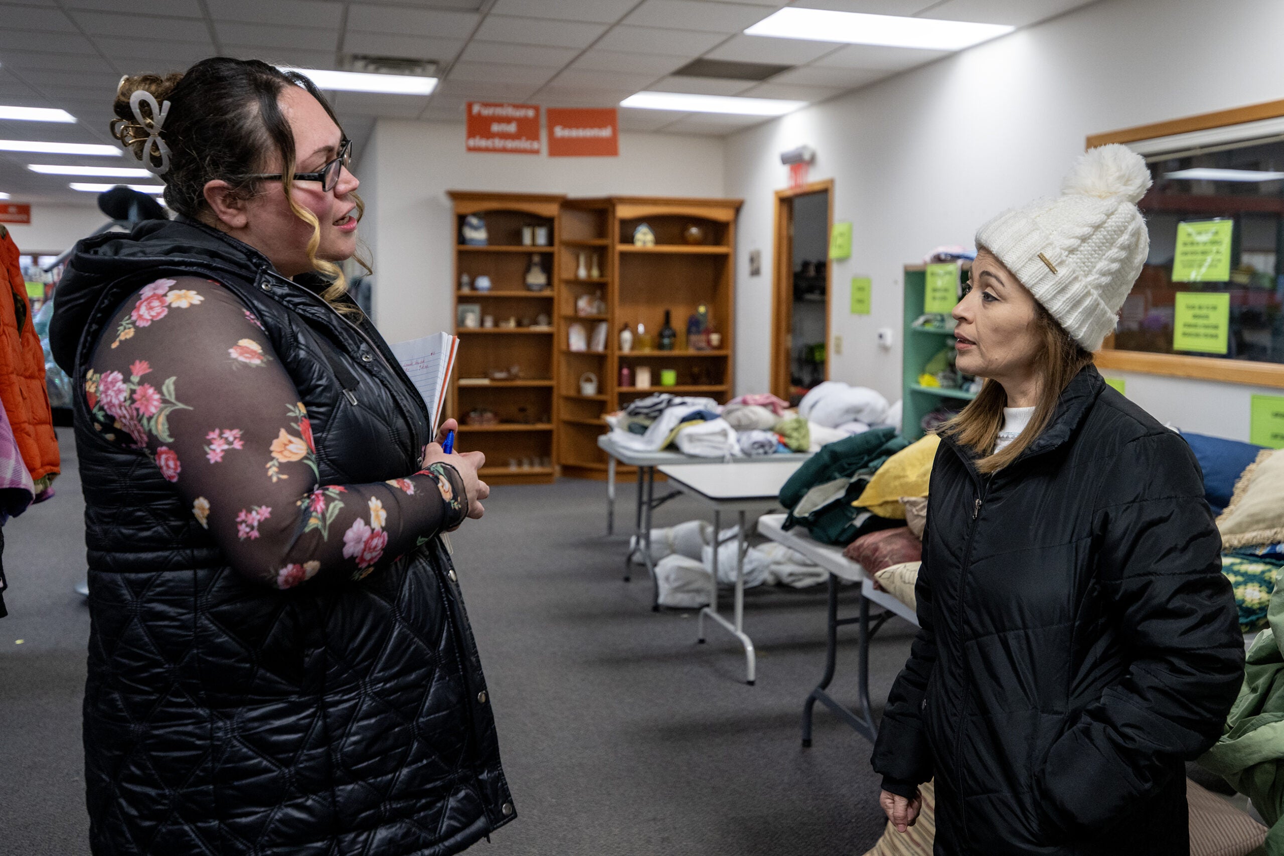 Two women wearing winter coats talk inside a room with shelves holding household items, folded blankets, and signs on the walls.
