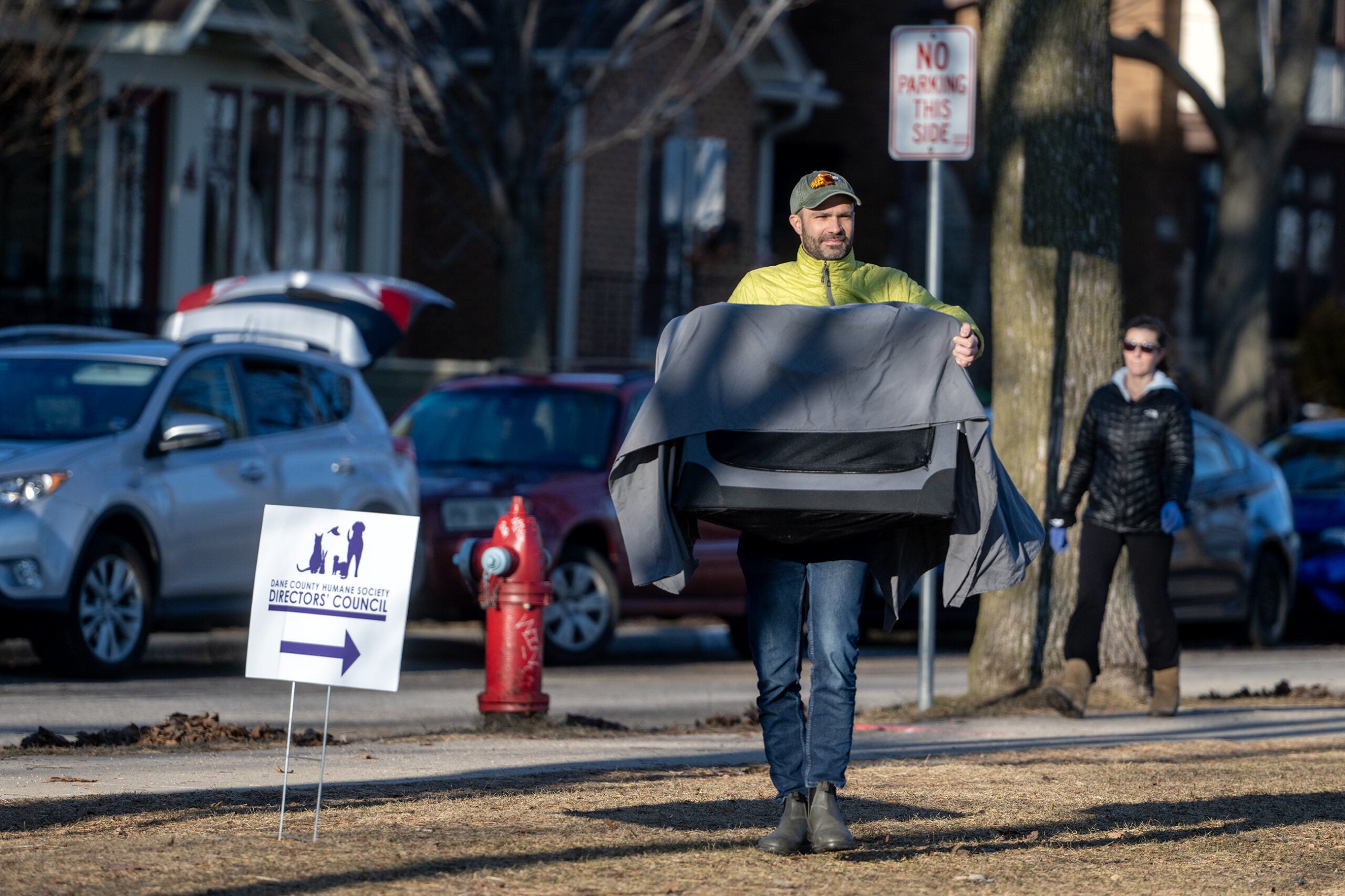 A man carries a folded chair across a grassy area near parked cars, with a street sign and a fire hydrant visible in the background. A woman stands nearby.