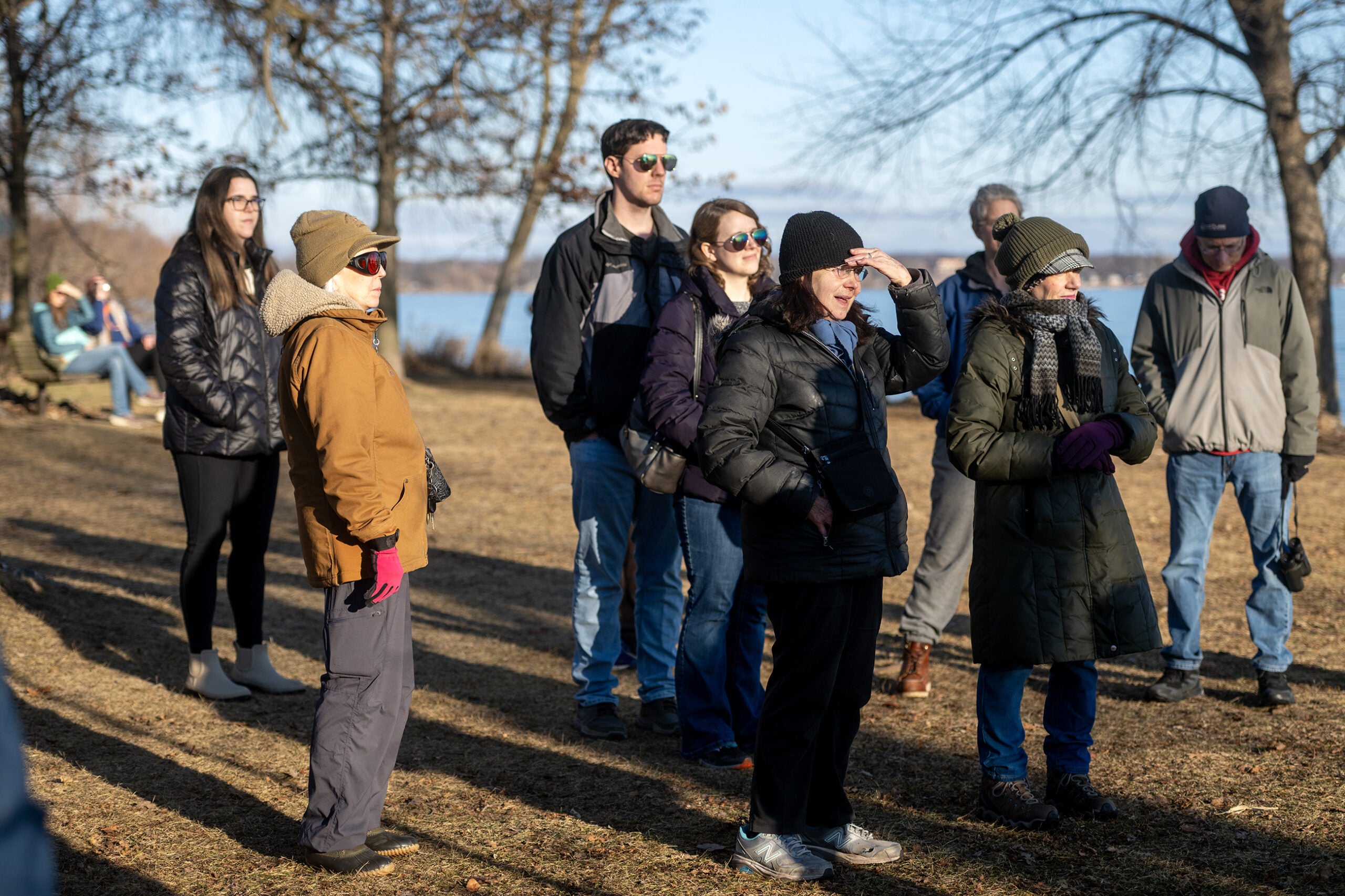 A group of people dressed in winter clothing stands outdoors on a sunny day near bare trees and a body of water, some looking into the distance.