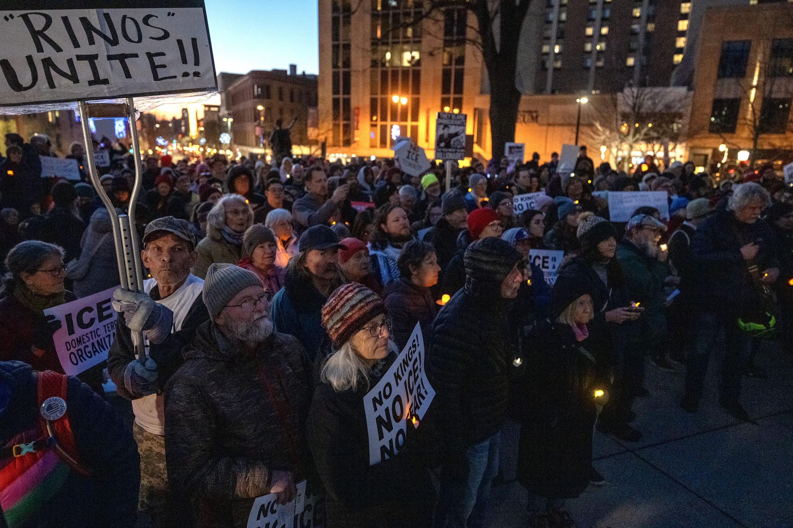 A large group of people gathers outdoors at dusk, holding signs with messages such as RINOS UNITE and NO ONE IS ABOVE THE LAW, during a protest or rally.