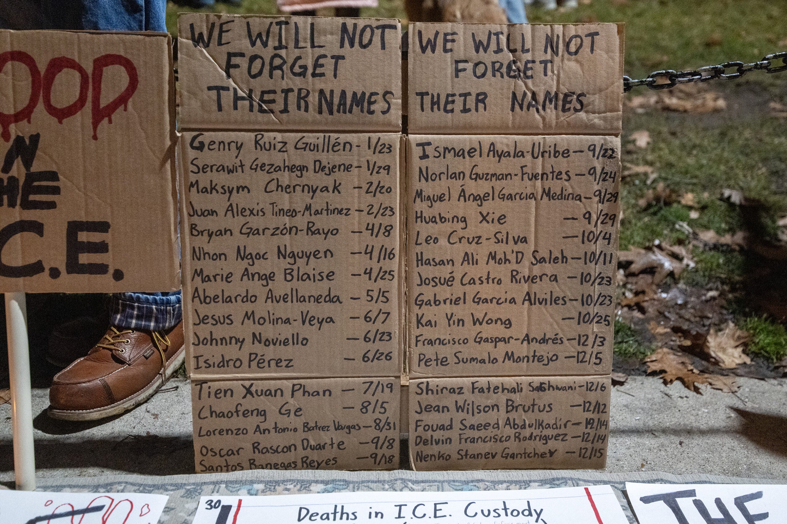 Cardboard signs list names and dates of individuals who died, with the heading We Will Not Forget Their Names, displayed outdoors on the ground.
