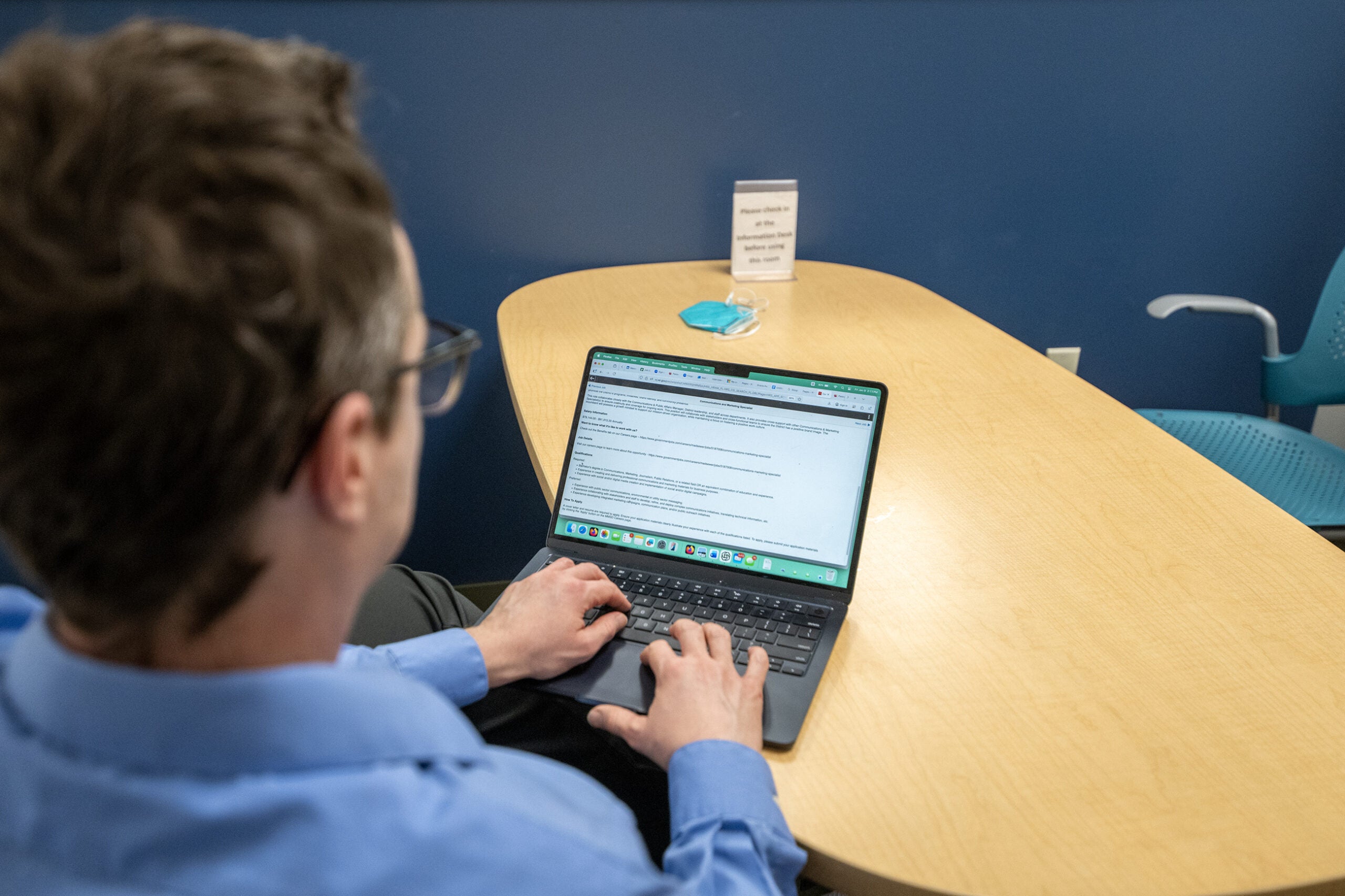 A person in a blue shirt types on a laptop at a table with a folded mask and a sign in the background.