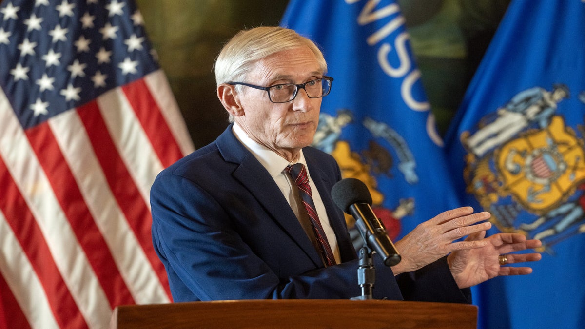 An older man in a suit speaks at a podium with a microphone, standing in front of American and Wisconsin state flags.