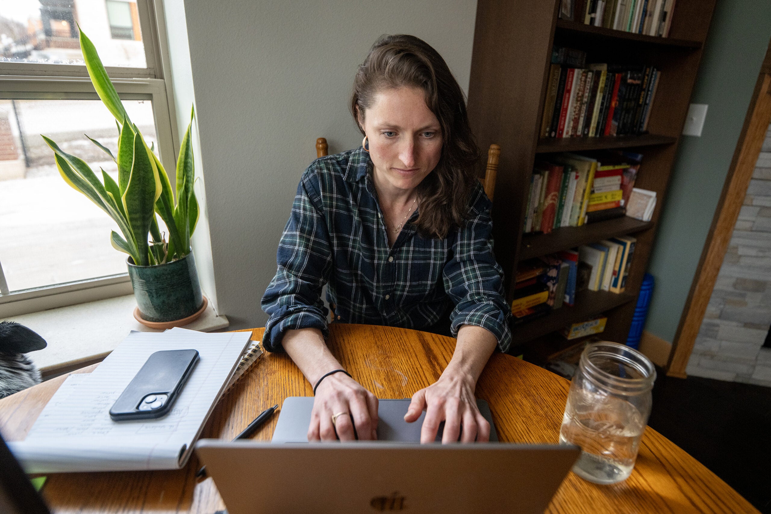 A person sits at a wooden table using a laptop, with a notebook, phone, glass jar of water, and a potted plant nearby. A bookshelf is in the background.