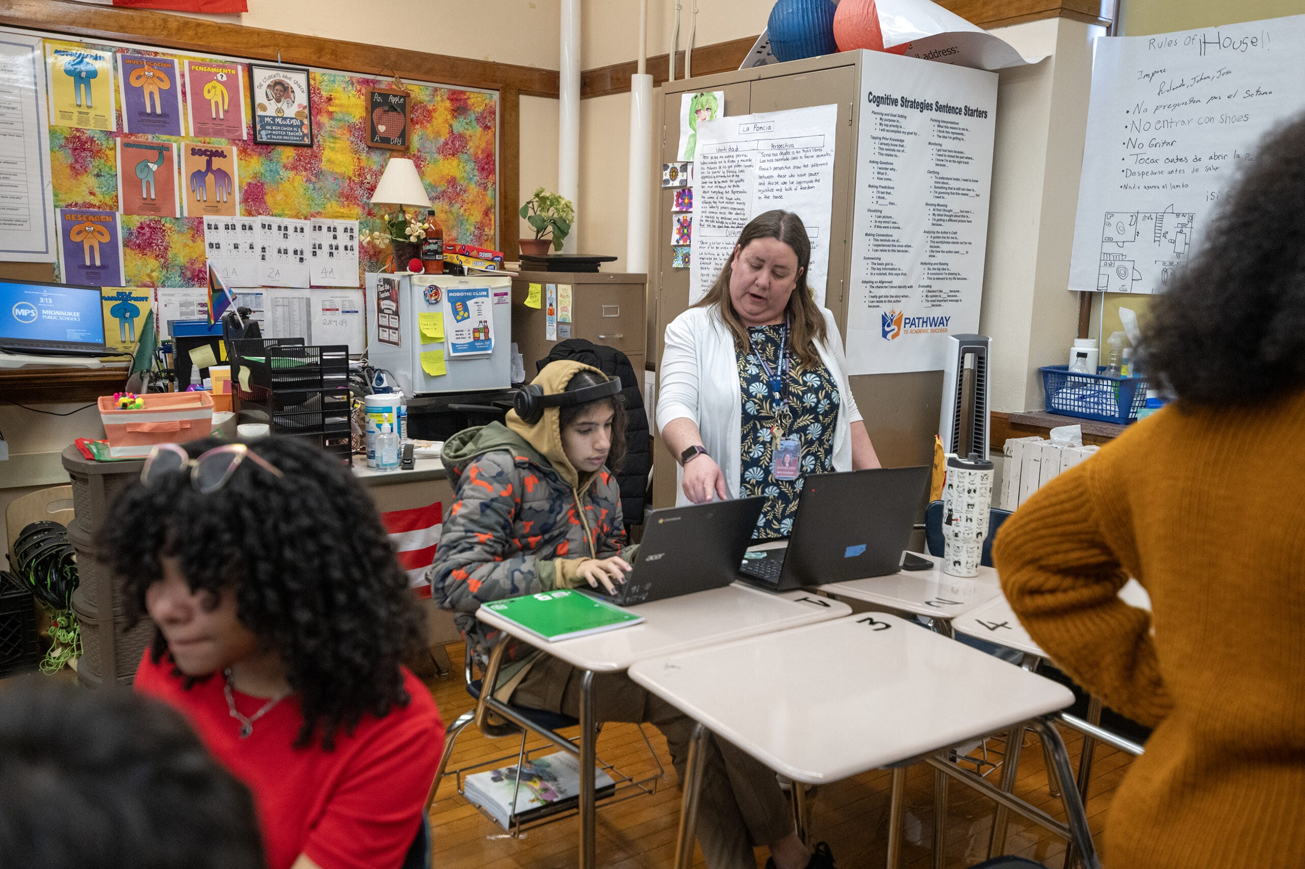 A teacher stands and points at a student’s laptop while several students work at their desks in a classroom.