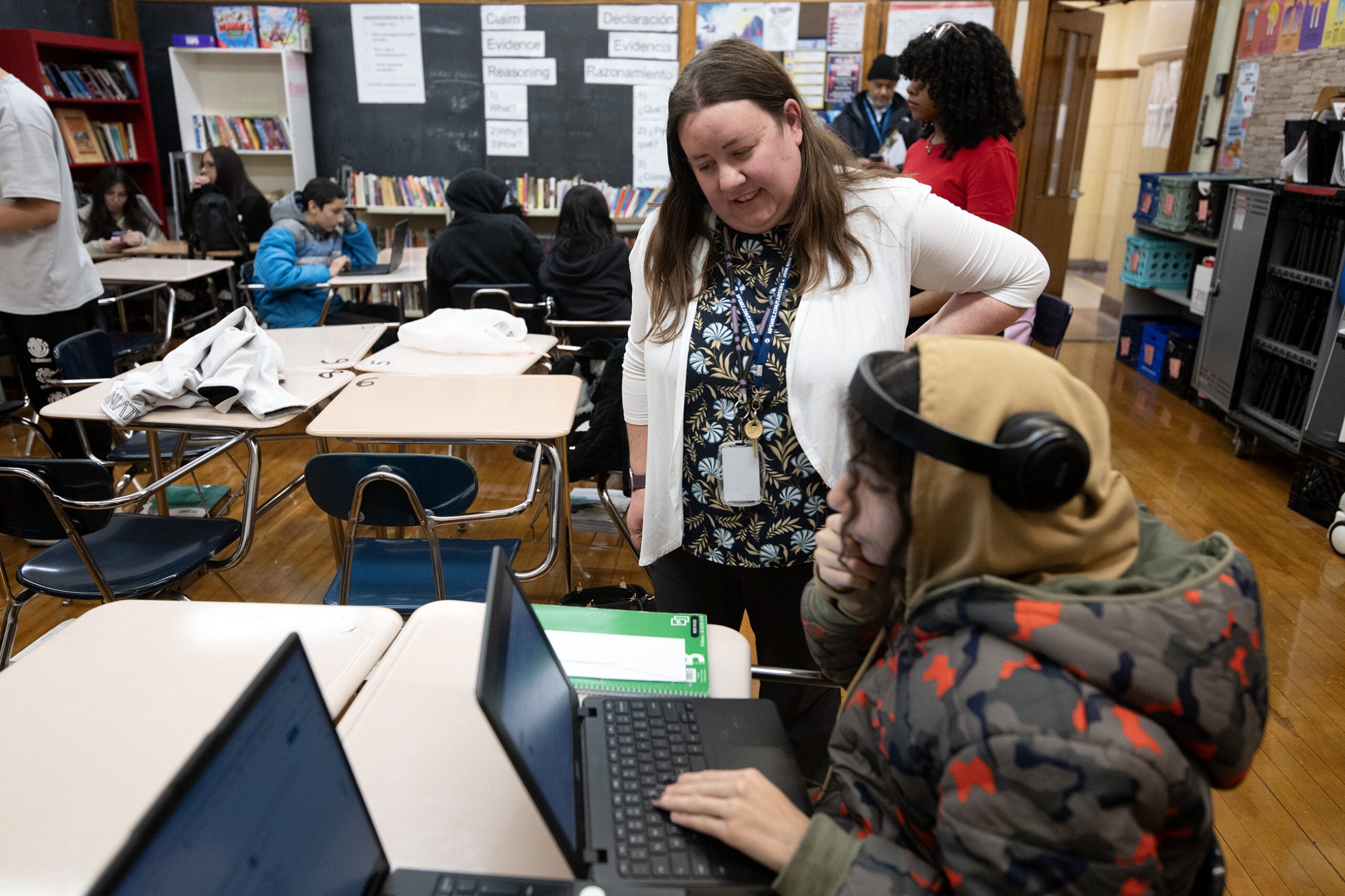A teacher stands next to a student wearing headphones who is using a laptop in a classroom with other students working at desks.