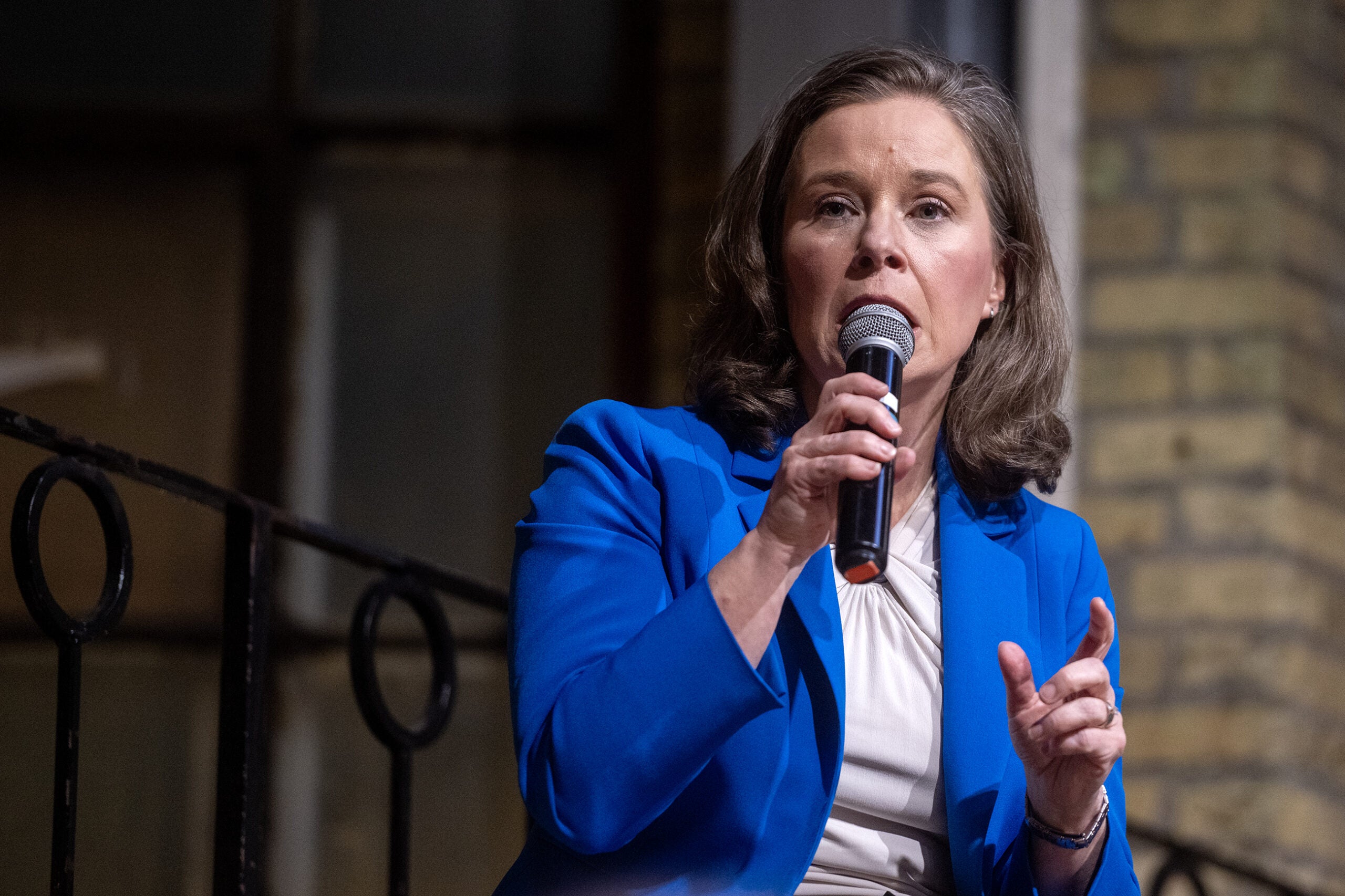 A woman in a blue blazer speaks into a microphone while gesturing with her hand, standing indoors near a railing and brick wall.