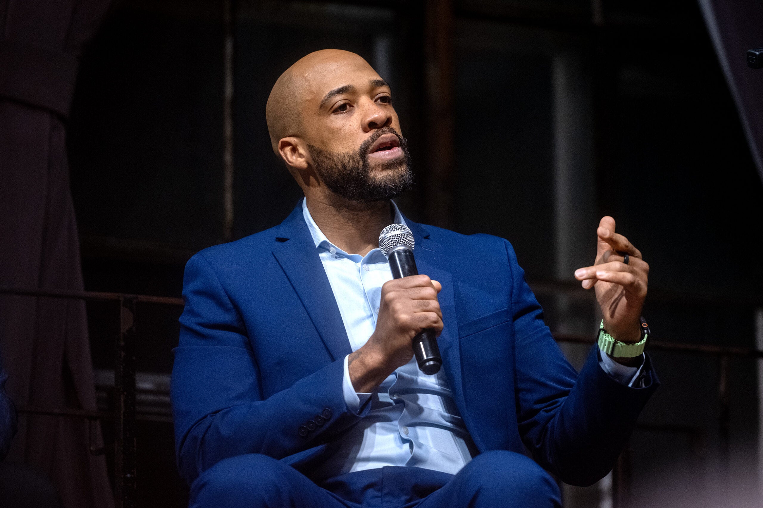 A man in a blue suit speaks into a microphone while gesturing with his hand during an indoor event.