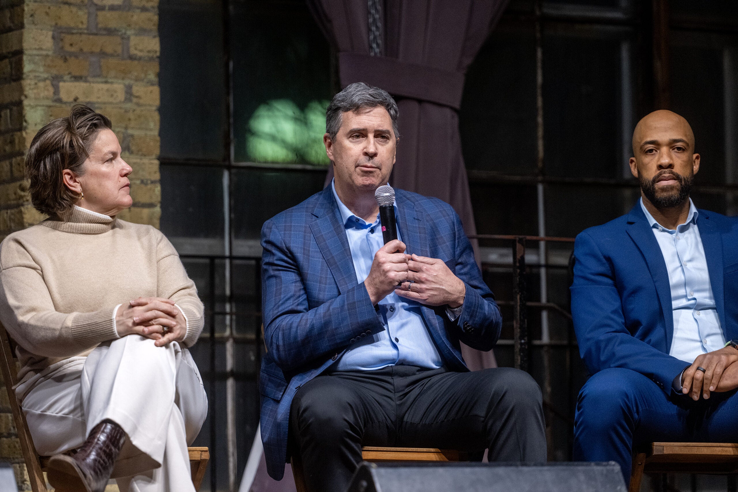 Three people sit on chairs; the person in the center, holding a microphone, speaks while the others listen. They are dressed in business attire against an indoor backdrop.