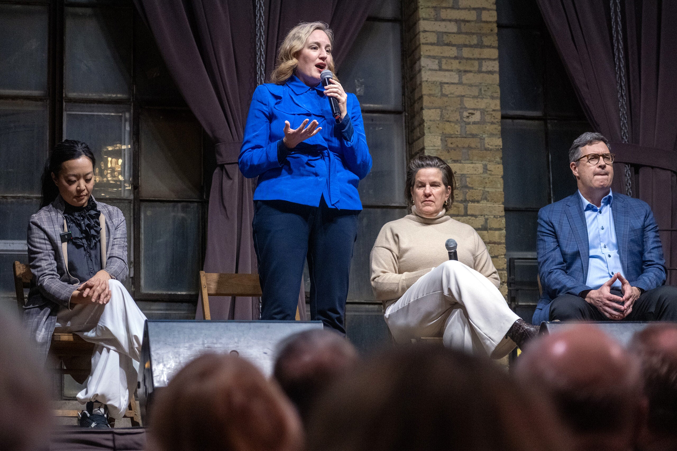 Four people sit on stage; the woman in blue stands holding a microphone and speaking, while the others sit with neutral expressions. Audience heads are visible in the foreground.