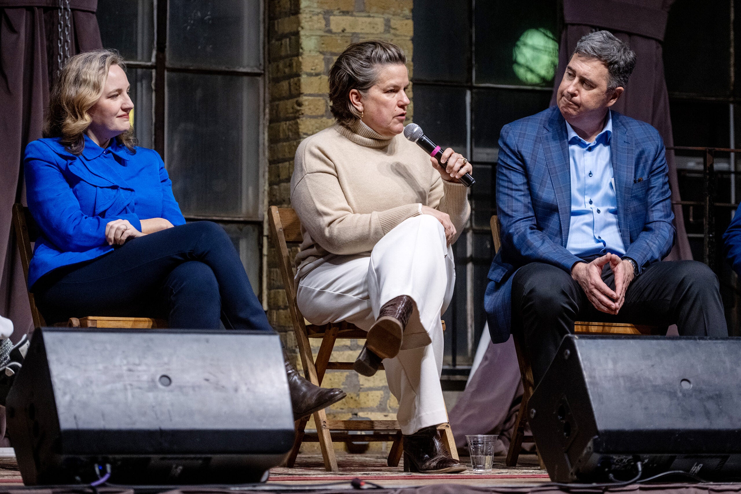 Three people sit on stage, two listening while the person in the center speaks into a microphone during a panel discussion.
