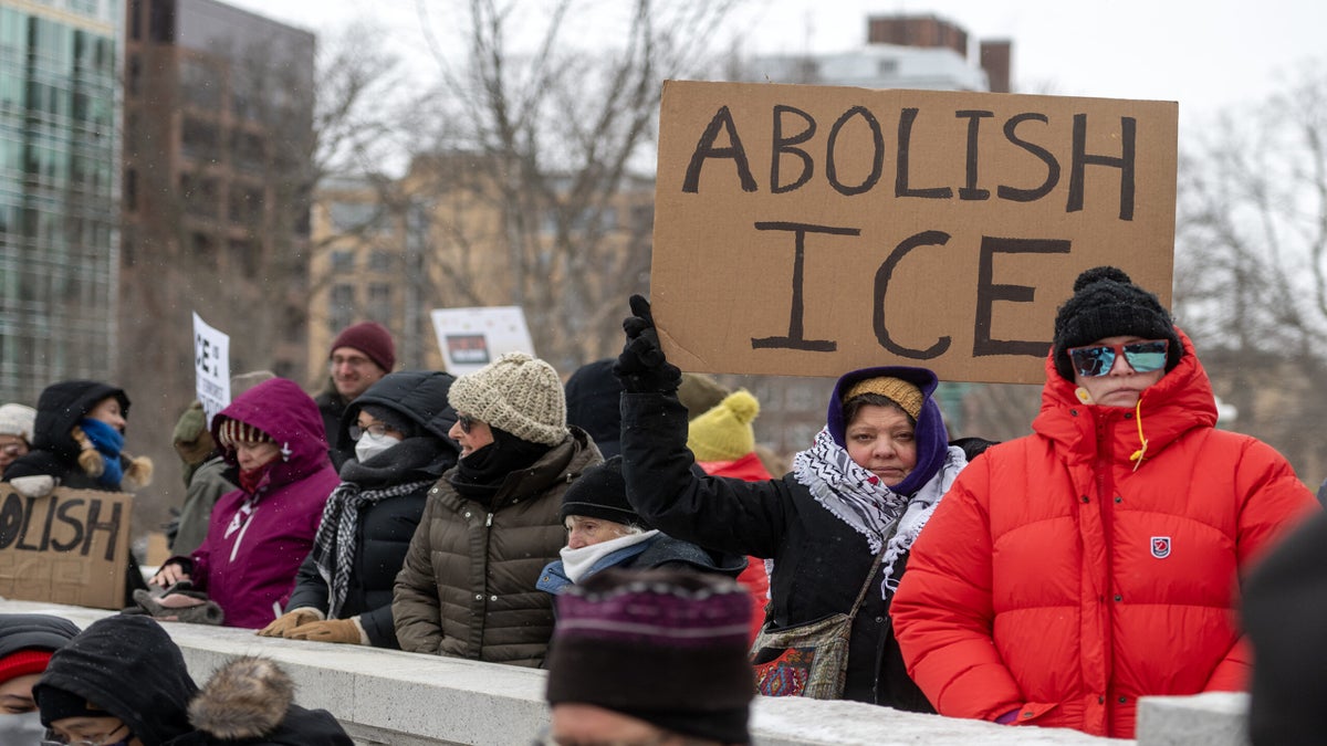 A group of people at an outdoor protest, one holding a large cardboard sign that reads ABOLISH ICE, with others dressed in winter clothing.