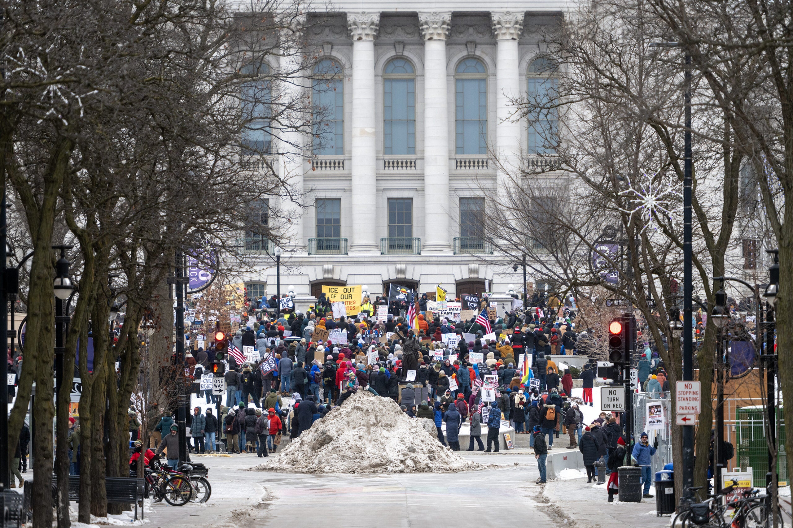 A large crowd of people holding signs gathers in front of a government building on a winter day; snow is piled in the street in the foreground.