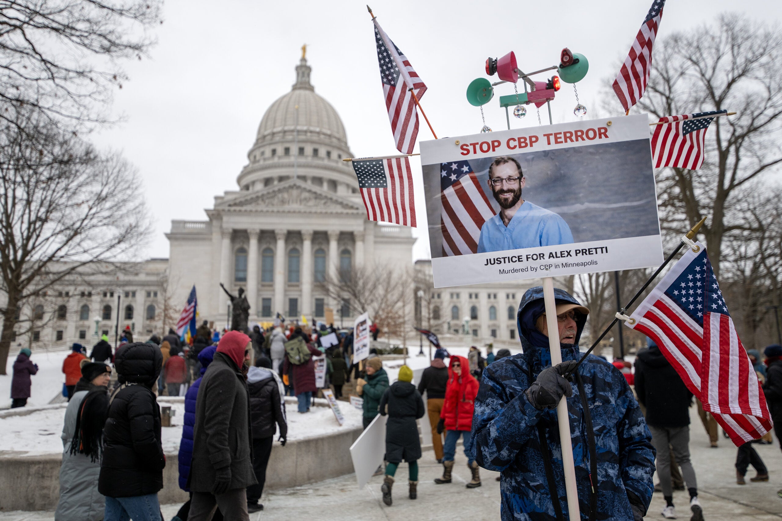 A person holds a sign with American flags and a photo in front of a government building during a winter protest; several people are gathered in the background.