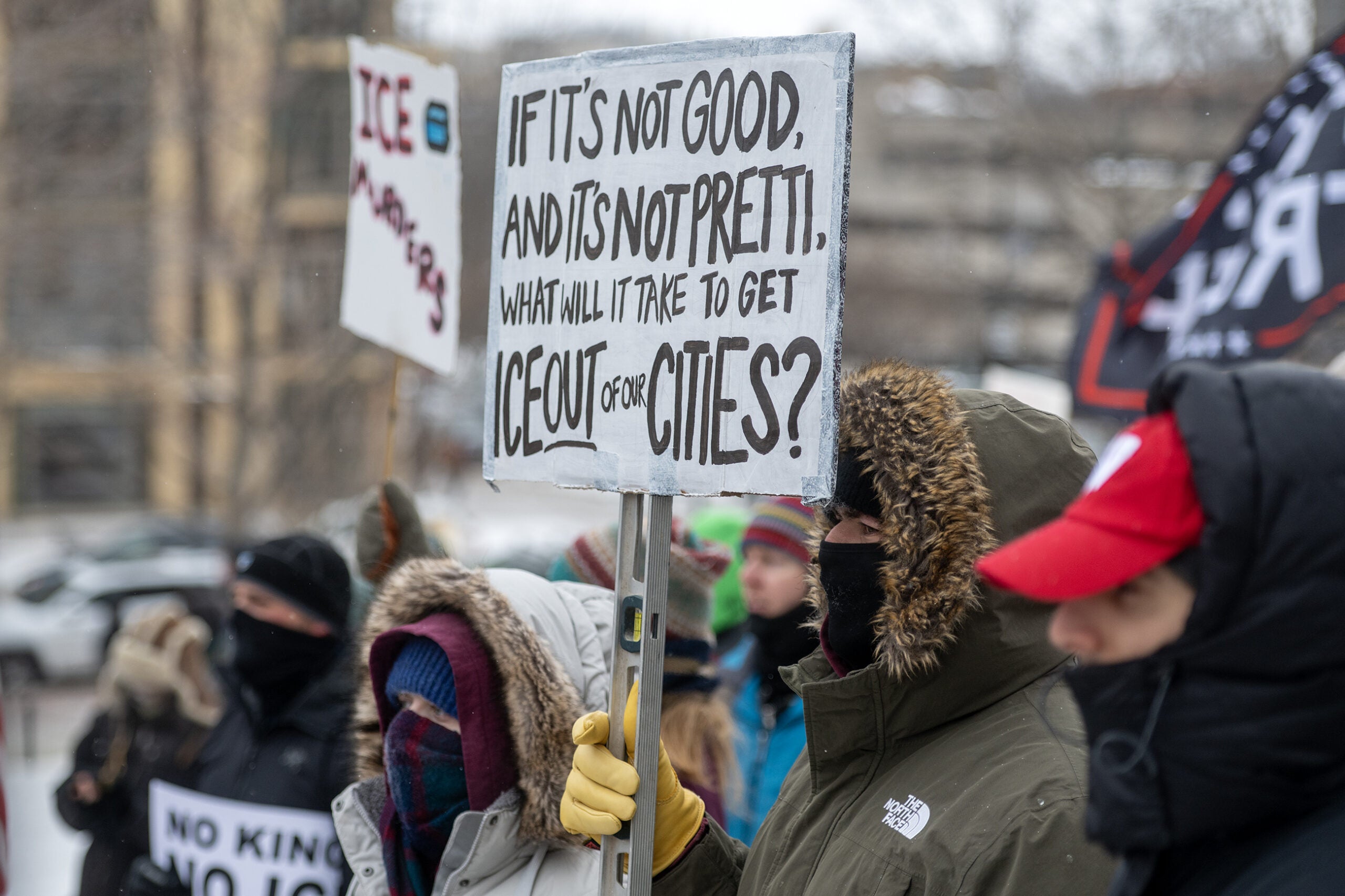 A group of bundled-up protesters stand outdoors in winter, holding signs, including one that reads, If it’s not good, and it’s not pretty, what will it take to get ICE out of our cities?.