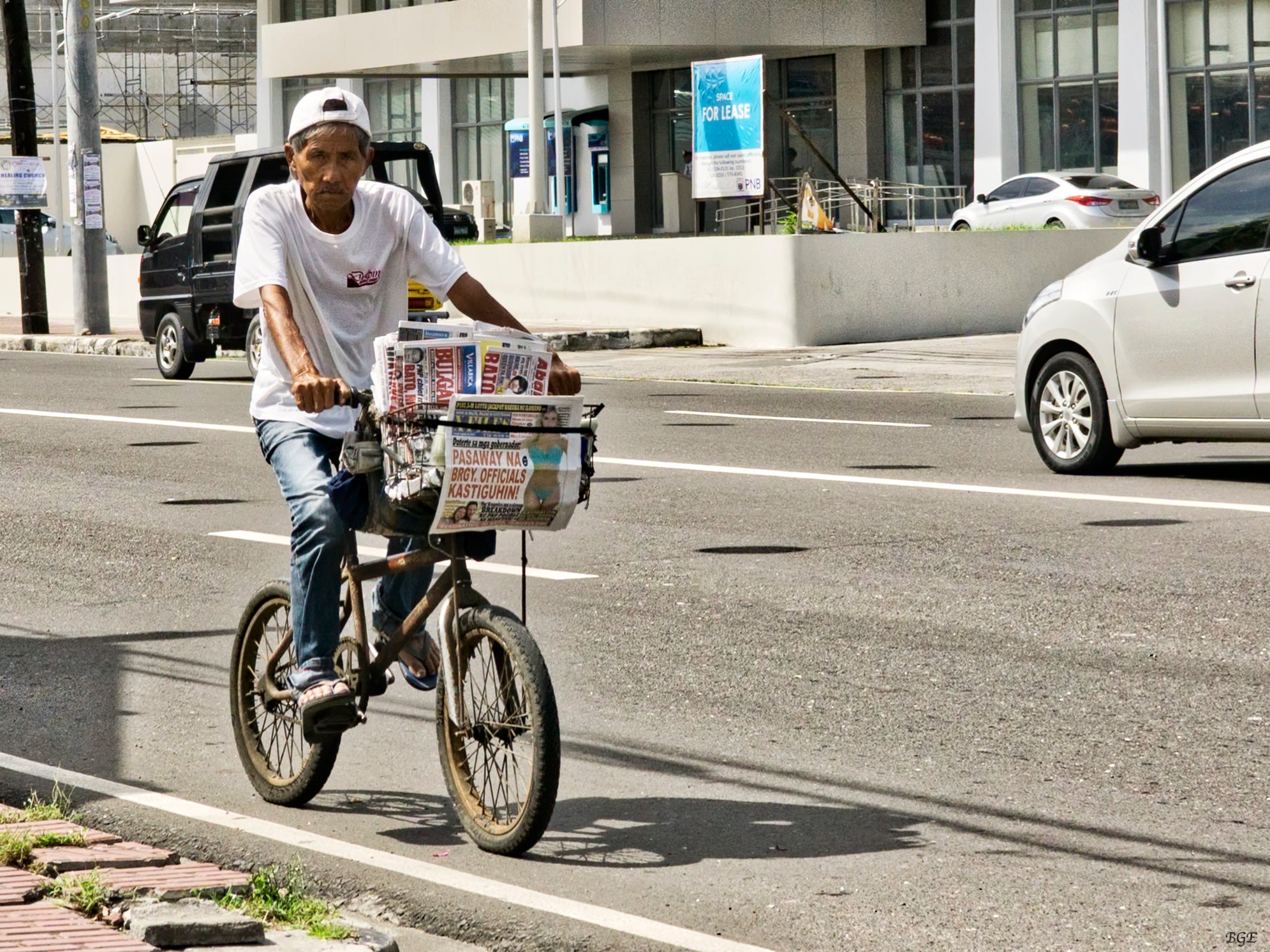 An elderly man rides a bicycle with a basket full of newspapers on a city street during the day.