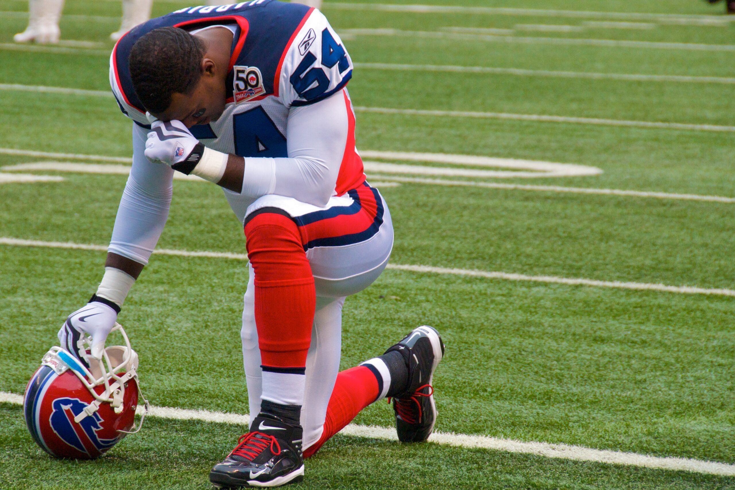 A football player wearing a white, red, and blue uniform kneels on one knee on the field, holding a helmet with his head bowed.