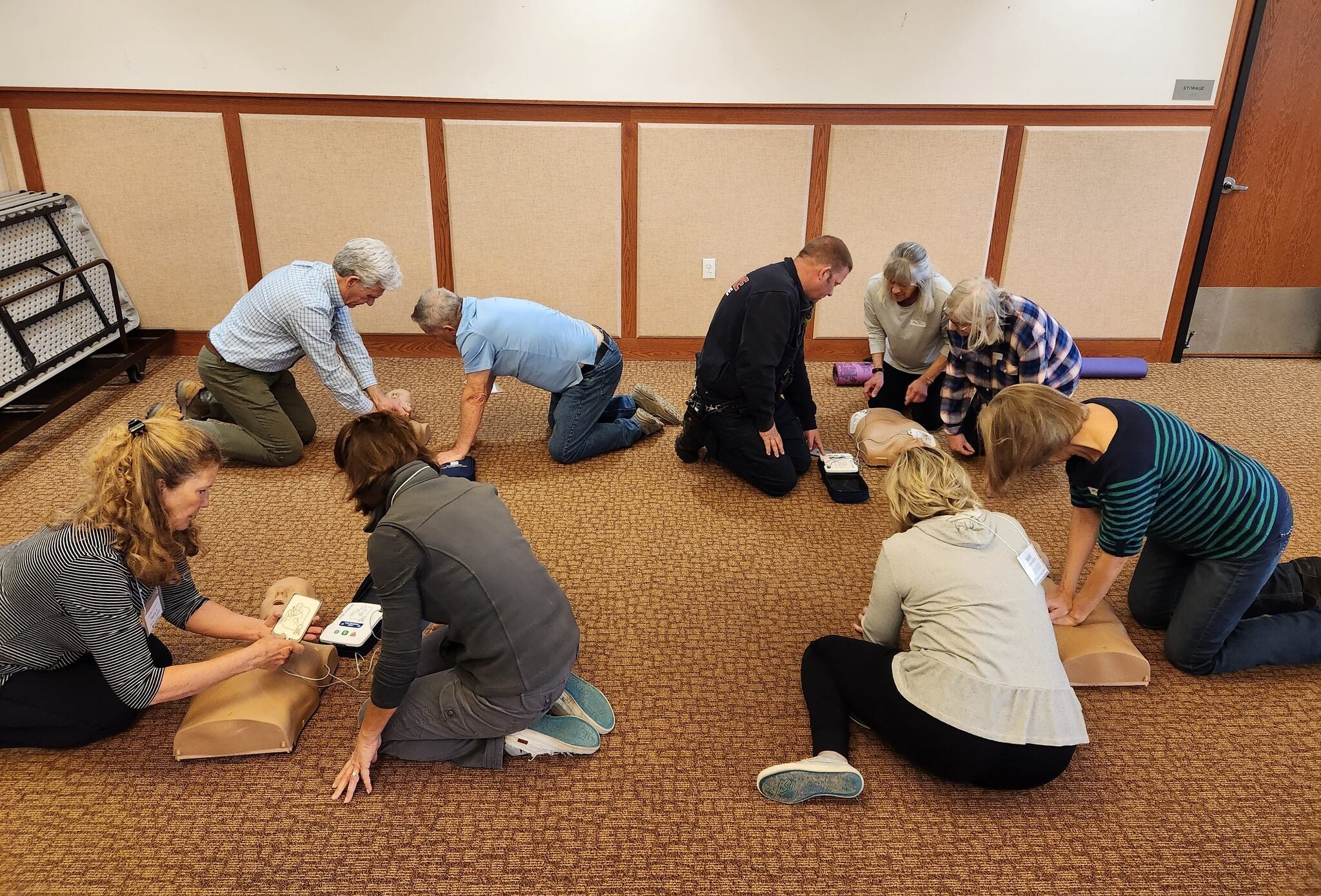 A group of adults kneeling on the floor practice CPR techniques on training mannequins during a class in a carpeted room.