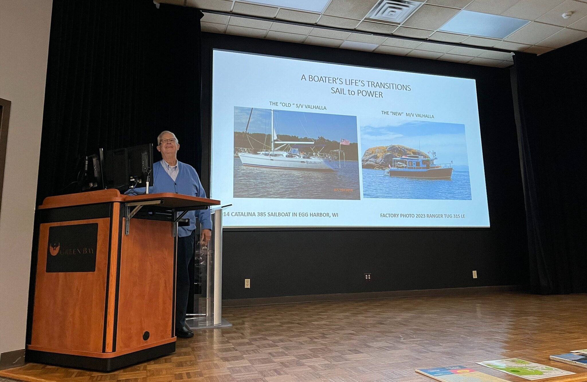 A man stands at a podium giving a presentation titled A Boaters Lifes Transitions: Sail to Power, with images of two boats displayed on a screen behind him.