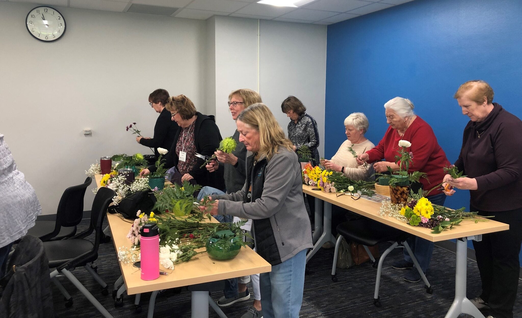 A group of people stand at tables in a classroom arranging flowers and greenery, with supplies and finished arrangements visible. A clock and blue wall are in the background.