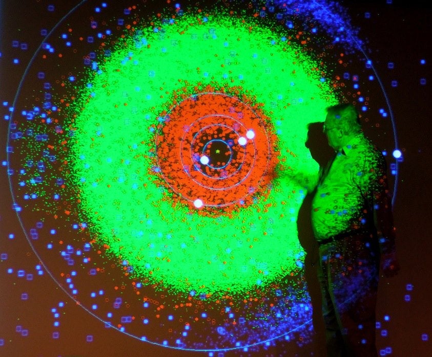 A person stands in front of a colorful projected diagram showing orbits and dense clusters of data points in red, green, and blue.