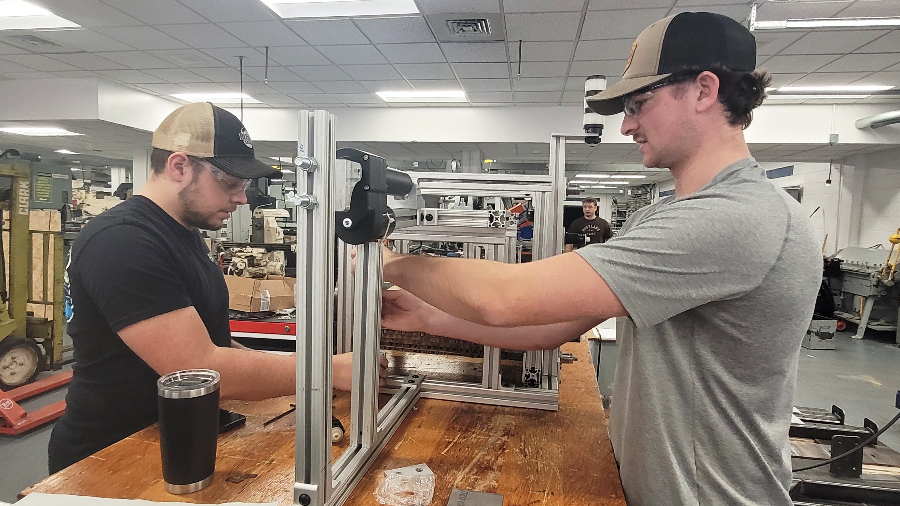 Two people in baseball caps work together assembling a metal frame structure on a table in a workshop, with tools and equipment visible in the background.