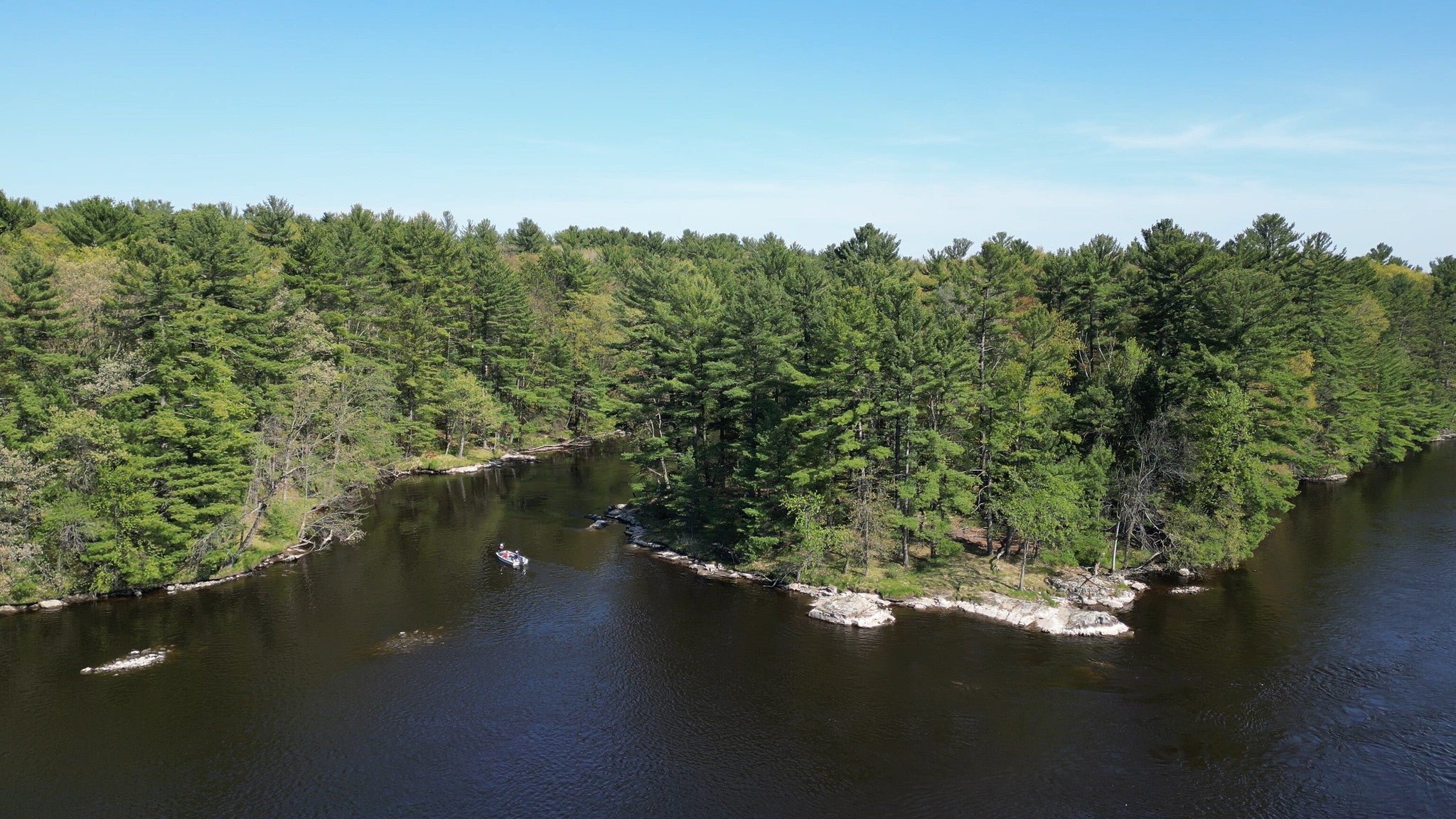 A river curves around a tree-covered island, with several kayakers paddling on the water under a clear blue sky.