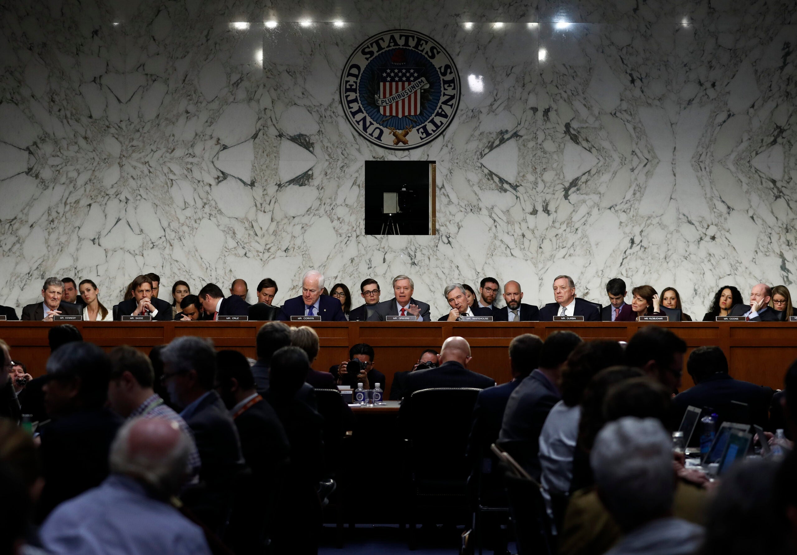 A congressional hearing in session, with officials seated at a long desk facing an audience, under a large United States Senate emblem on a marble wall.