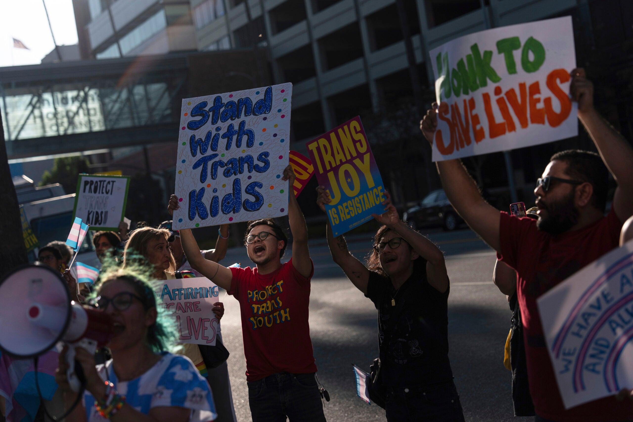 A group of people hold signs and chant at a protest in support of trans youth; signs read “Stand With Trans Kids” and “Trans Joy Is Resistance.”.