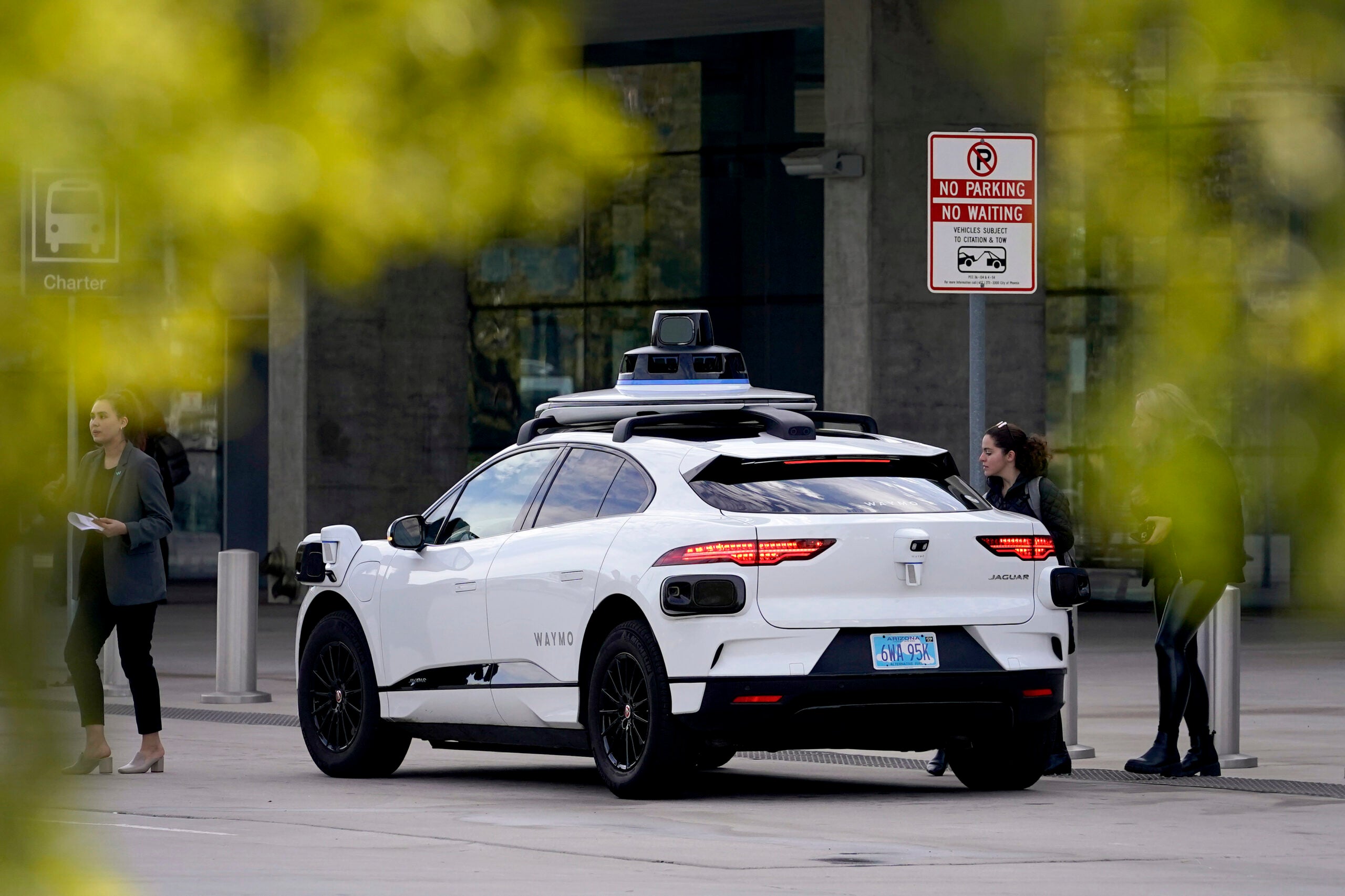 A white autonomous vehicle with sensors on the roof is parked near a sidewalk; two women stand nearby, and a no parking sign is visible in the background.