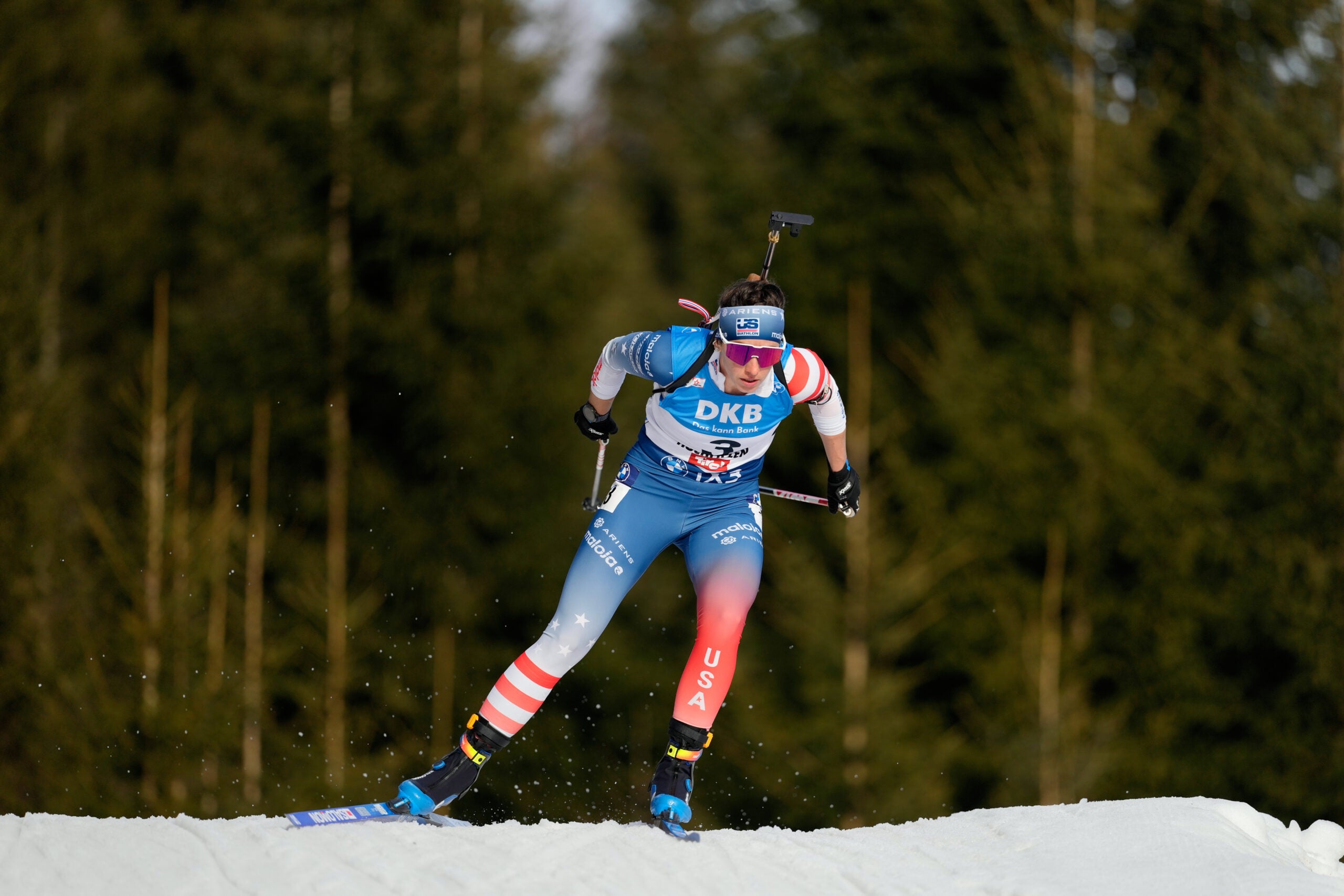 A biathlete wearing blue and red USA race gear skis on snow with a rifle on their back, against a background of trees.