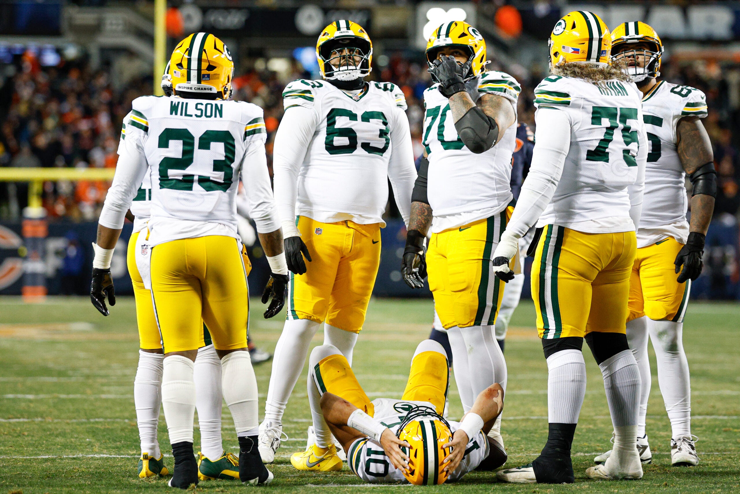 Green Bay Packers football players stand around a teammate lying on the field during a game, with stadium lights and spectators in the background.