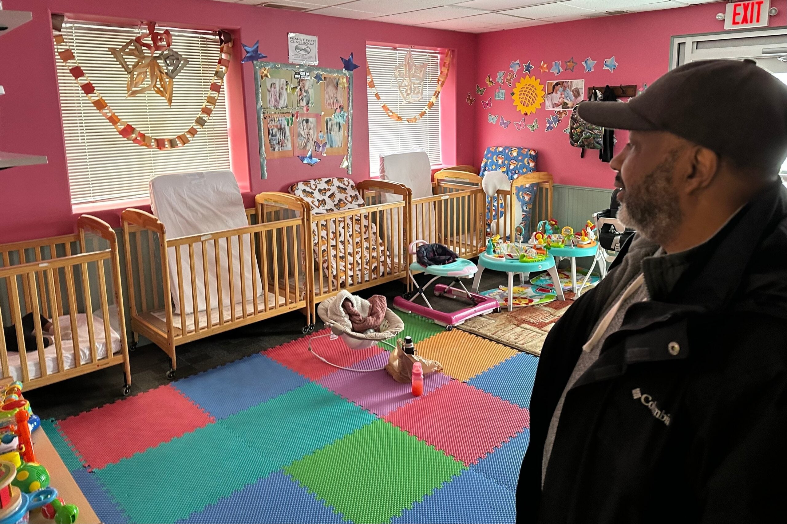 A man stands in a daycare room with colorful mats, empty cribs, baby toys, and decorations on pink walls.