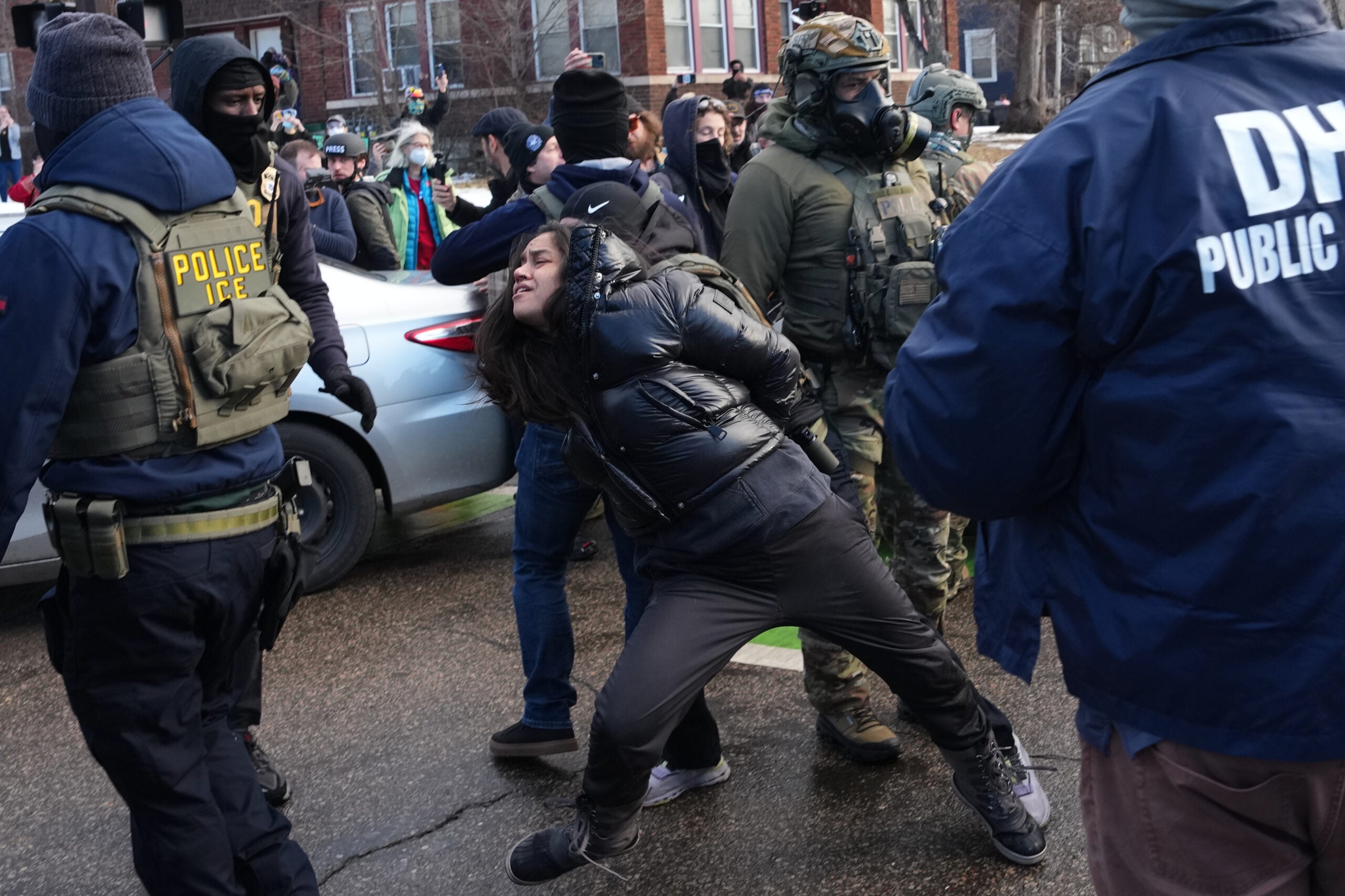 A person is being detained by law enforcement officers during a protest; several officers and bystanders are present in the background.