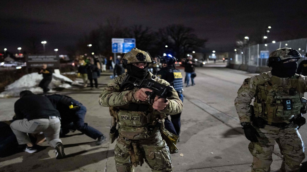 Armed police officers in tactical gear stand on a city street at night as several people are detained on the ground in the background.