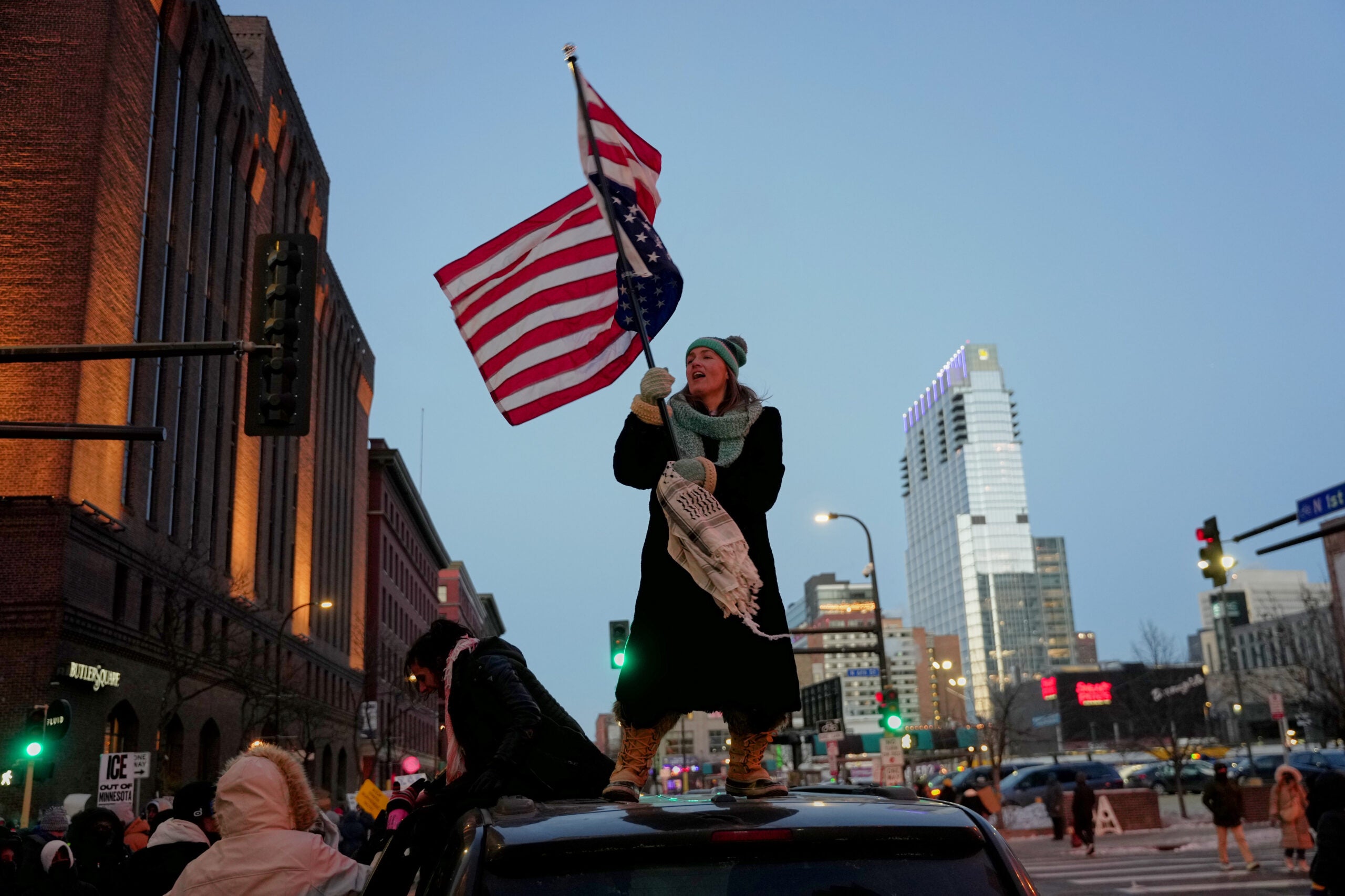 A person stands on top of a car holding an upside-down U.S. flag during a city street protest at dusk, surrounded by buildings and other demonstrators.