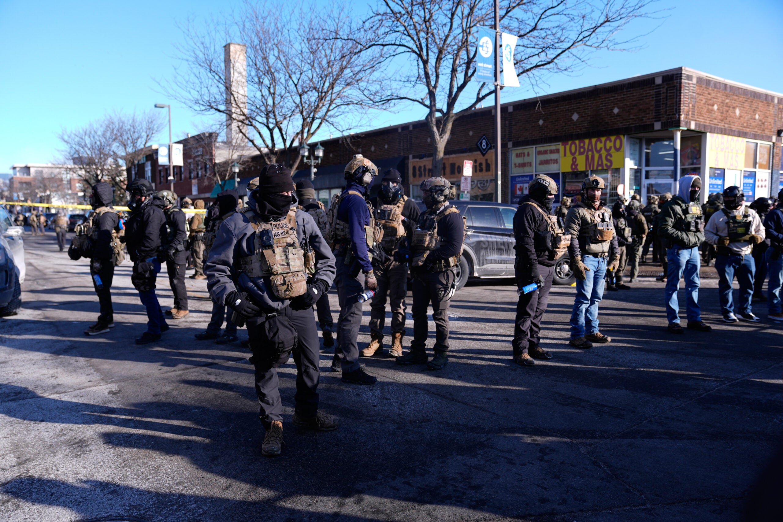 A group of law enforcement officers in tactical gear stand on a city street near stores, with police tape visible in the background.