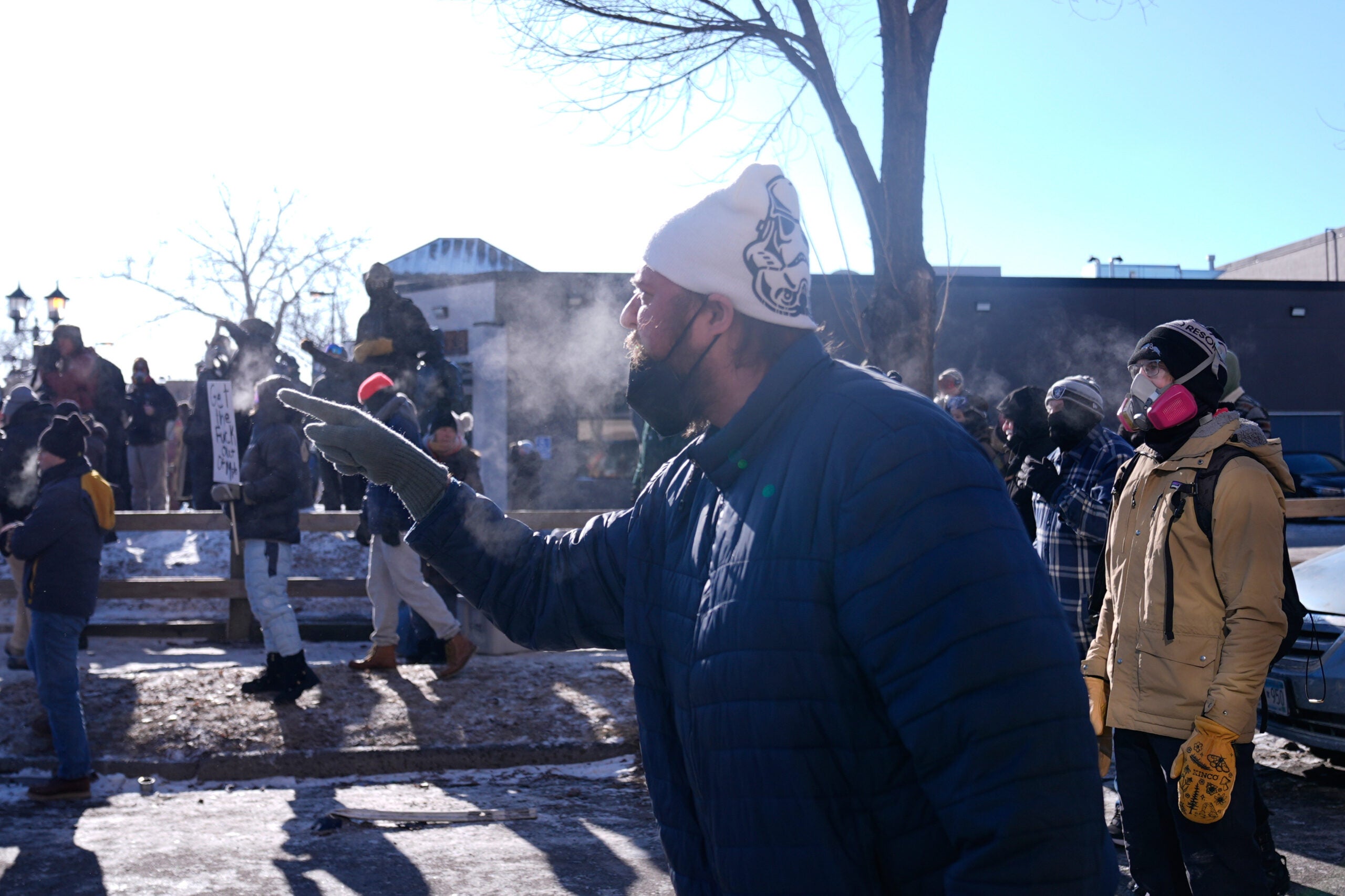 A person in a blue jacket and white beanie gestures and speaks outdoors on a cold day, surrounded by a group of people. Breath is visible due to the cold.