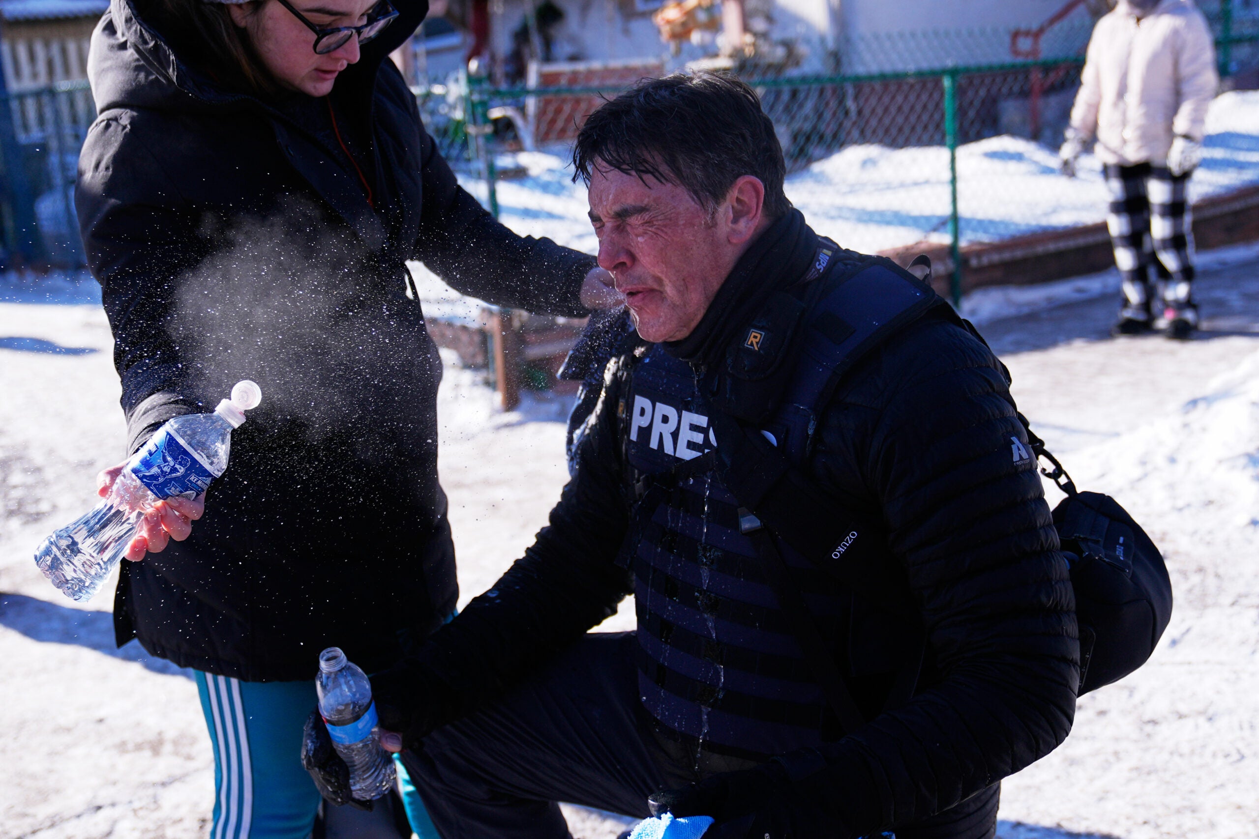 A person wearing a PRESS vest kneels on snowy ground while another person sprays water on his face from a bottle; both appear distressed and cold.